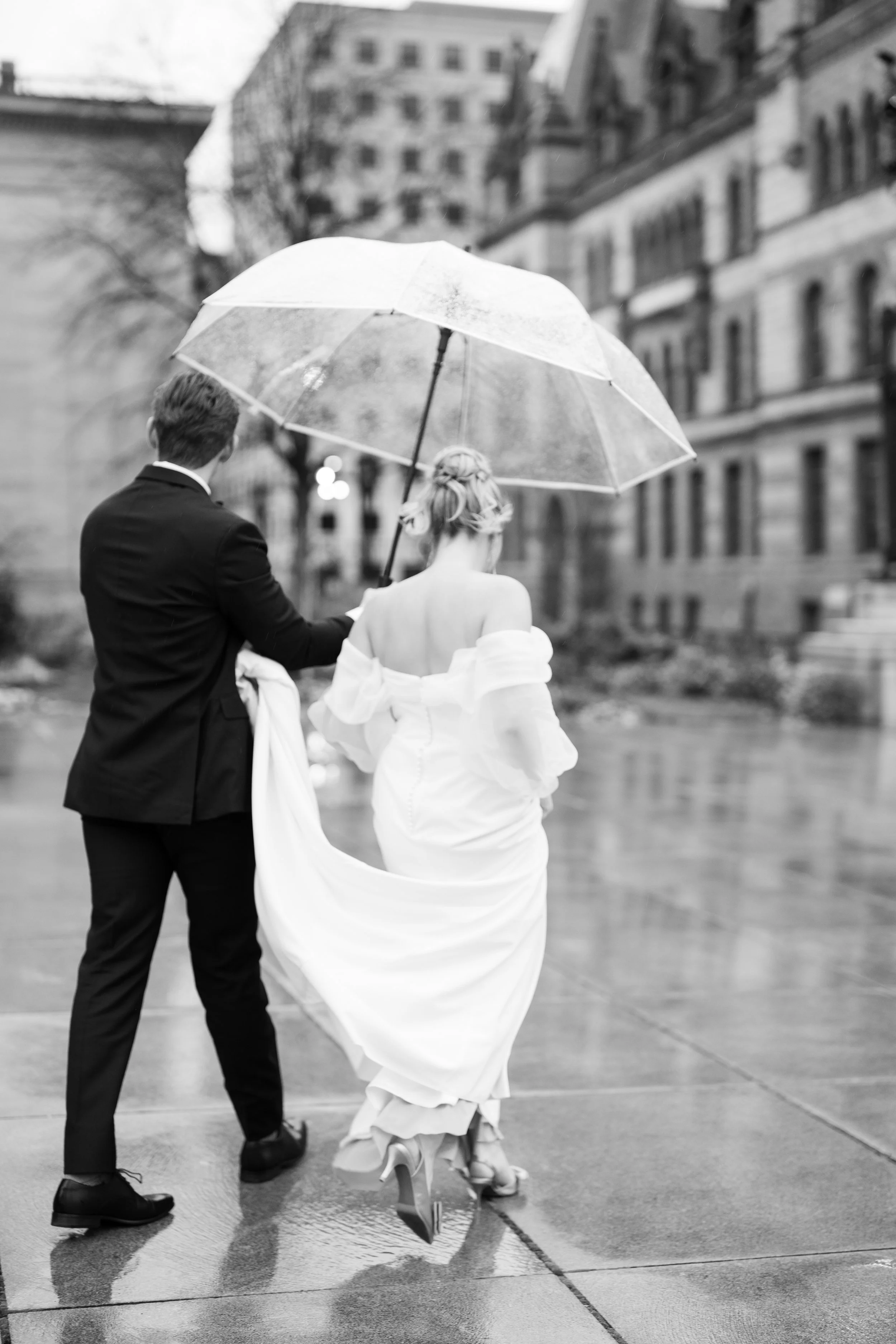 Black-and-white wedding portrait of a bride and groom walking beneath a clear umbrella in downtown Cincinnati on a rainy wedding day. Timeless, romantic wedding photography.