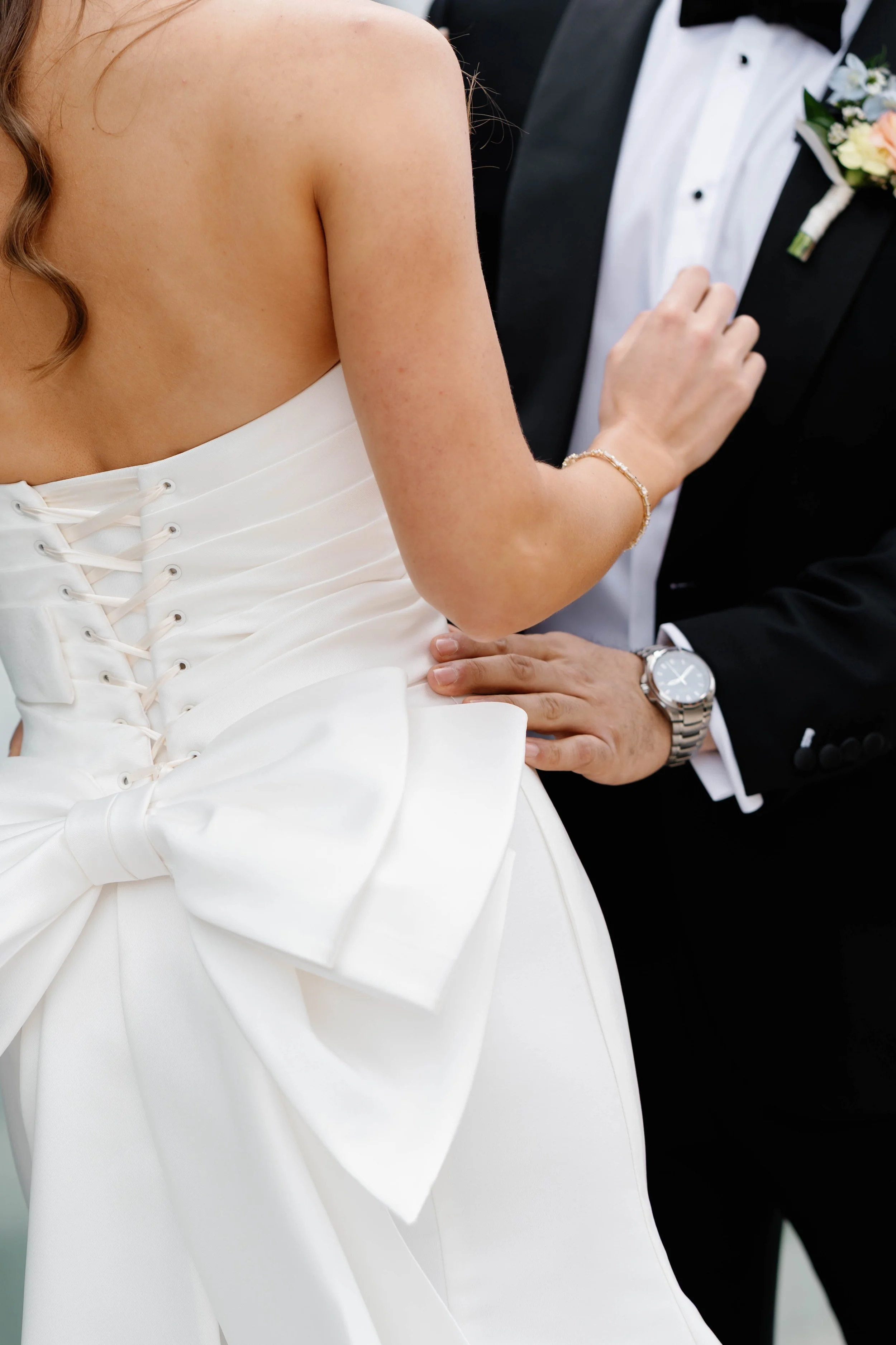 Close-up of a bride and groom, the bride in a white wedding dress with lace-up back, the groom in a black tuxedo with boutonniere and a watch, standing close with the bride touching the groom's chest.