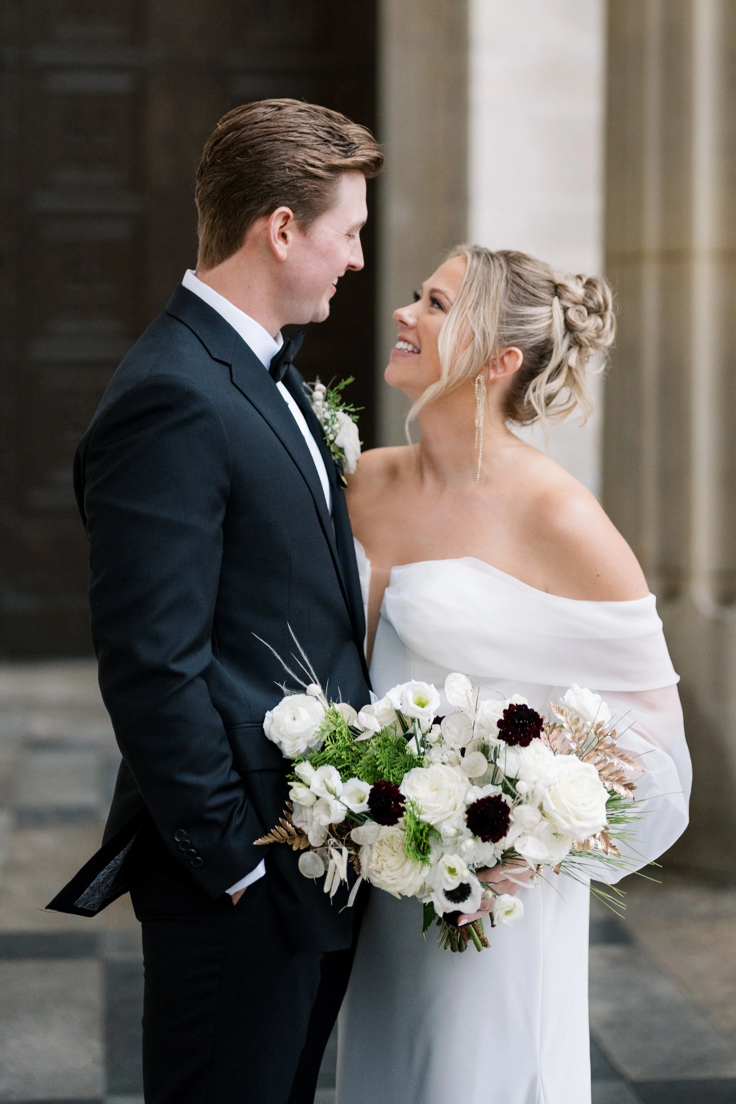 Bride and groom smile at one another while holding a white bridal bouquet during their wedding portraits.