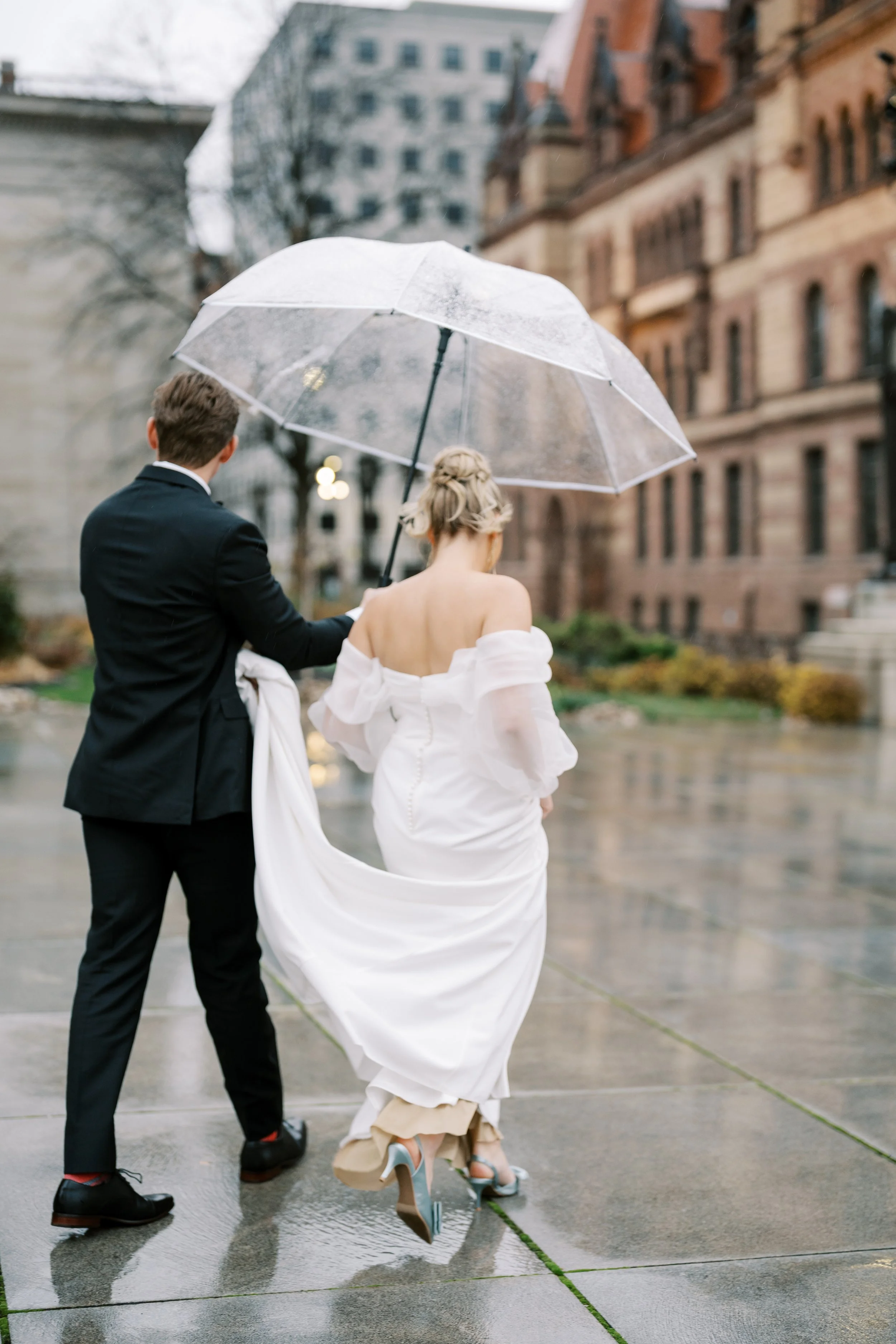 Bride and groom walking hand in hand beneath a clear umbrella during a rainy portrait session outside the Cincinnati Club.