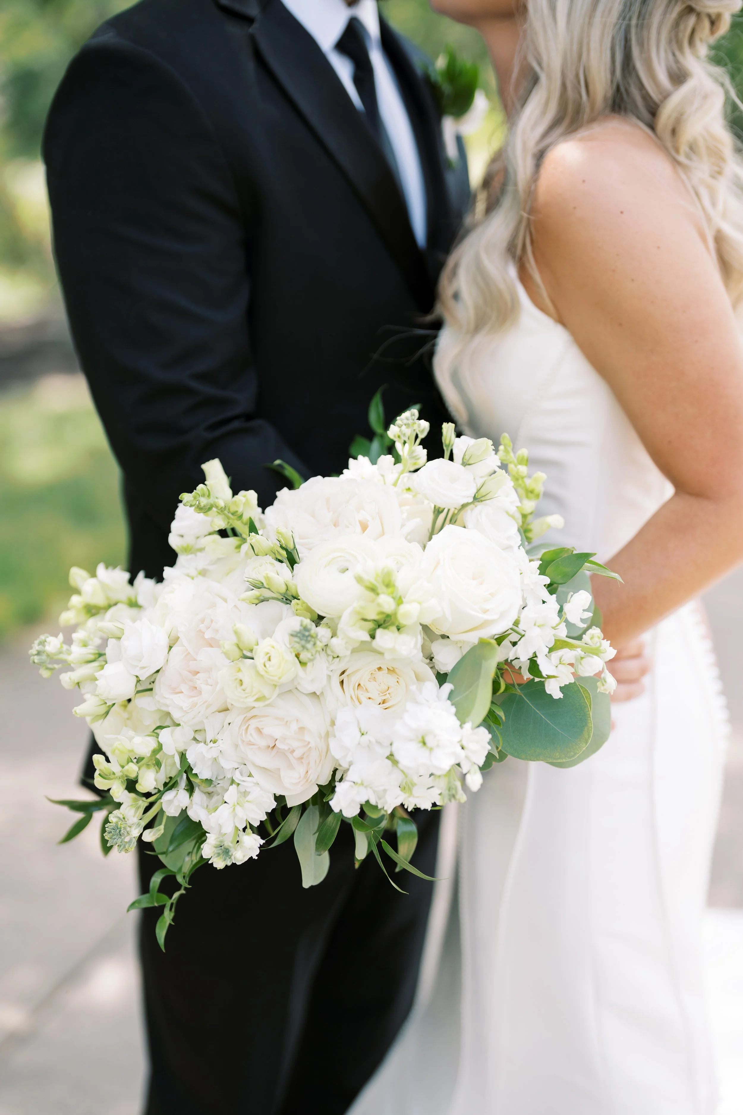 A bride and groom close together, holding a bouquet of white flowers, outdoors on a sunny day.