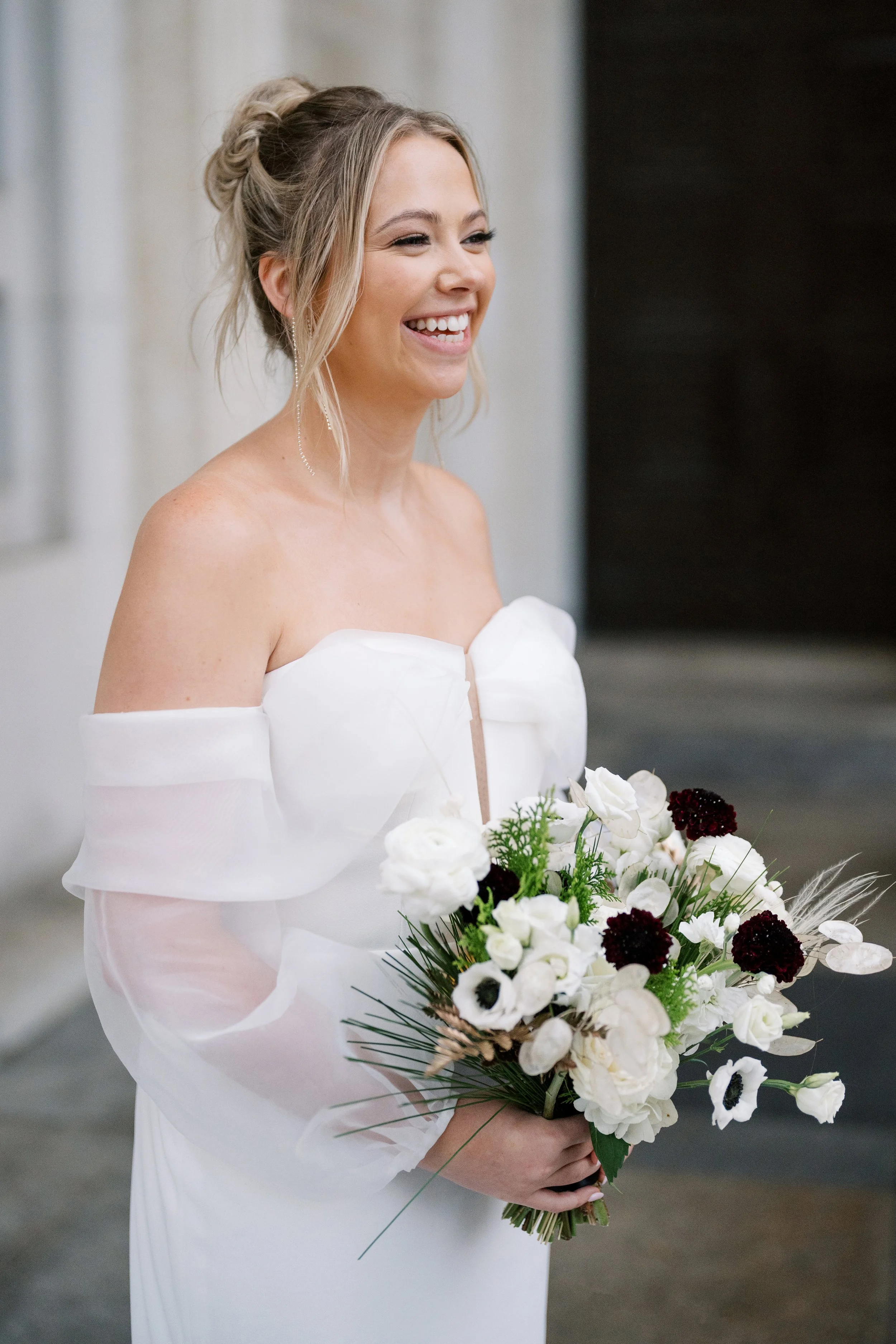 Bride smiles while holding her wedding bouquet in a modern off-the-shoulder wedding gown.