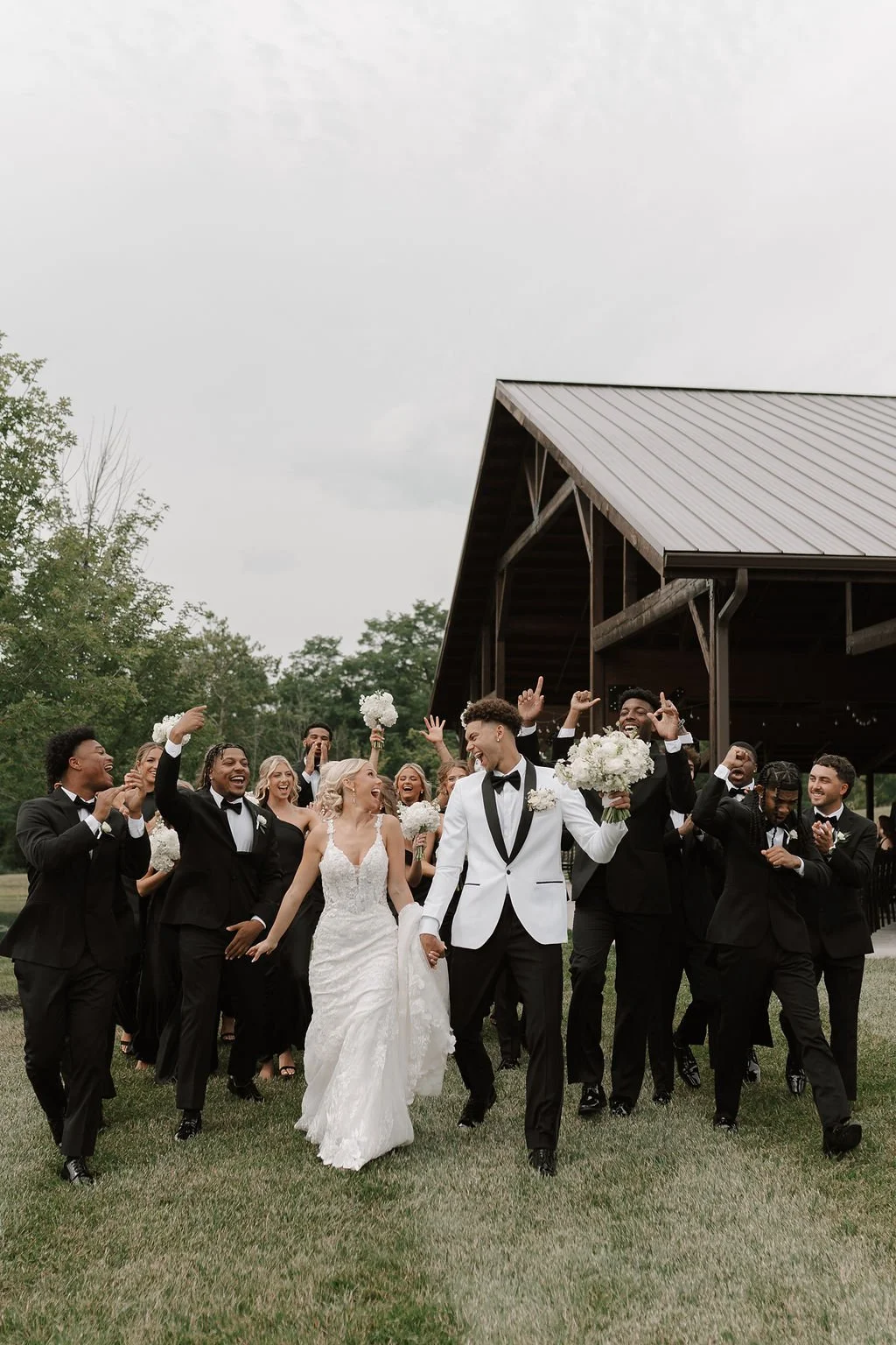 Bride and groom celebrate with their wedding party as they walk across the lawn outside a rustic wedding venue, surrounded by cheers and excitement.
