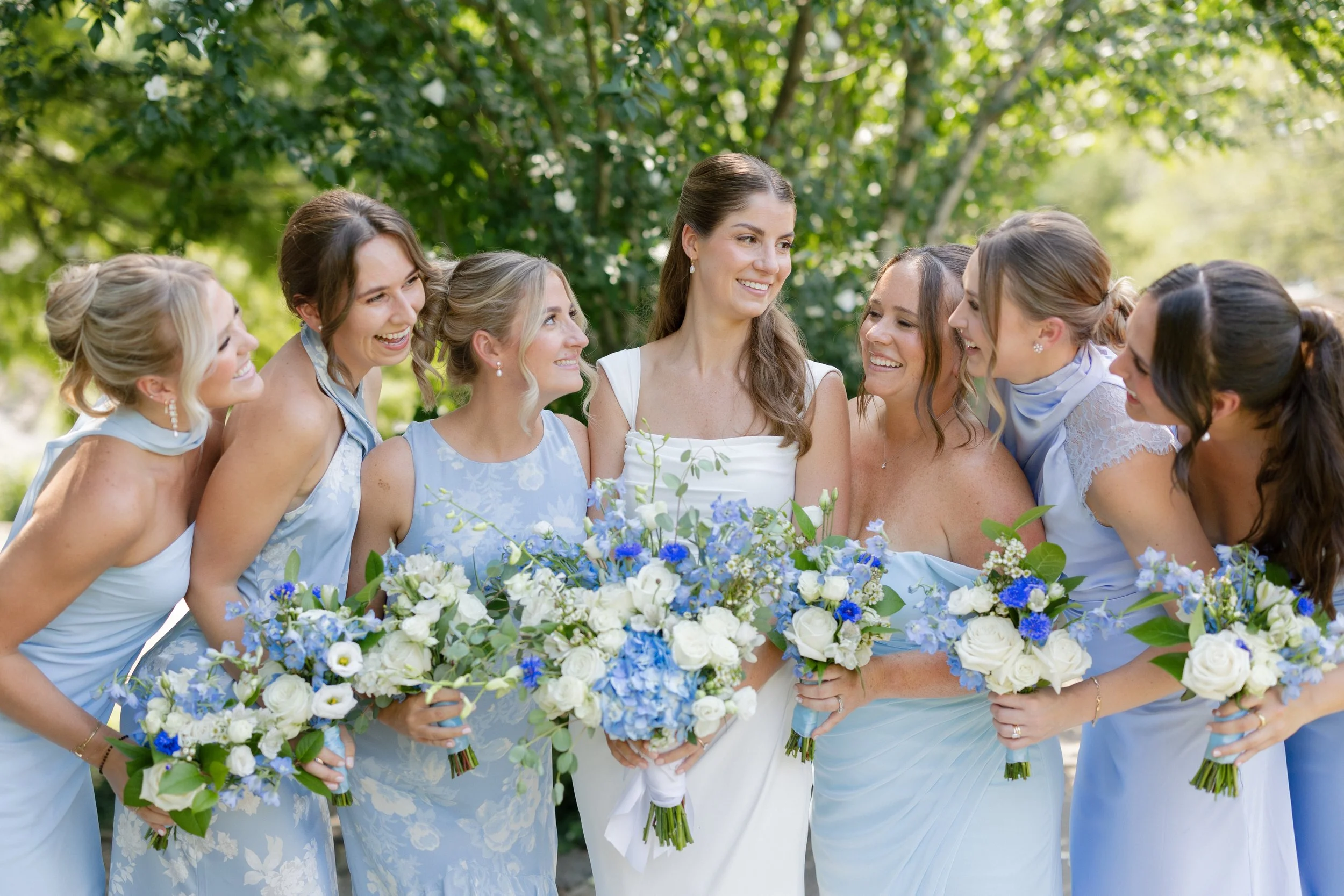 Bride laughing with her bridesmaids while holding blue and white floral bouquets during outdoor wedding portraits.