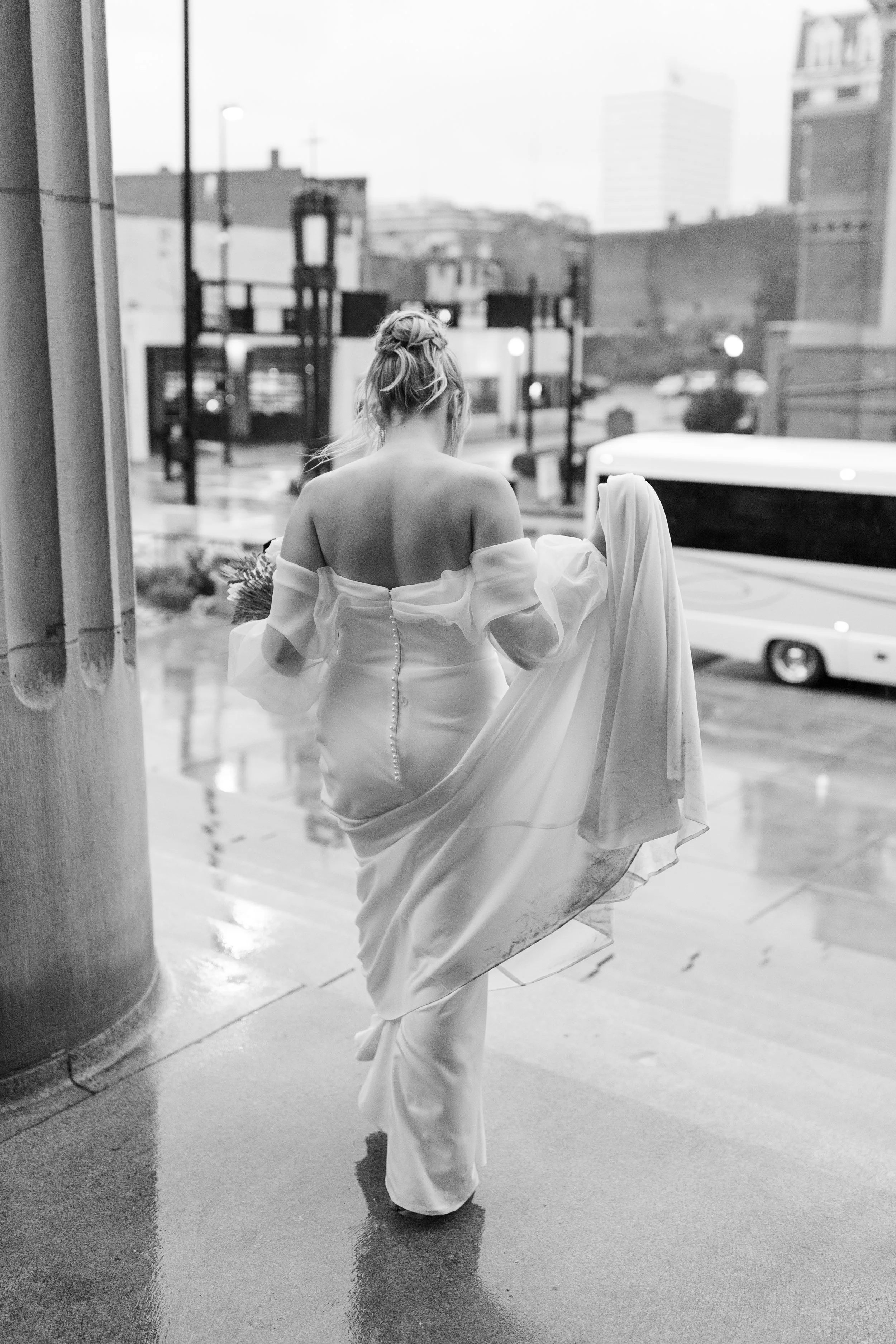 Bride walks beneath a clear umbrella through a rainy city street, her veil flowing behind her in a romantic black-and-white portrait.