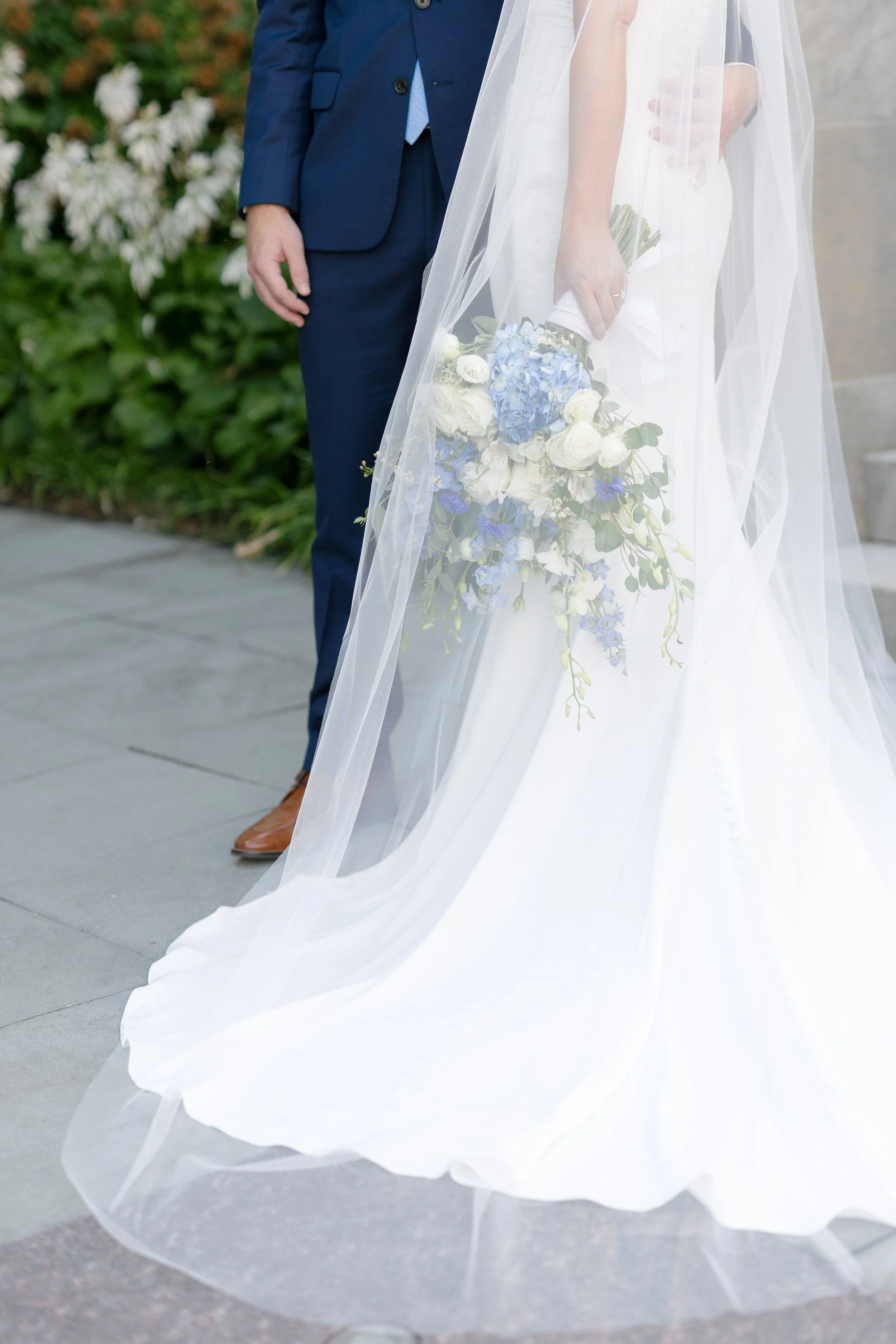 Bride holds a cascading bouquet of white and blue flowers beside her wedding gown and veil.
