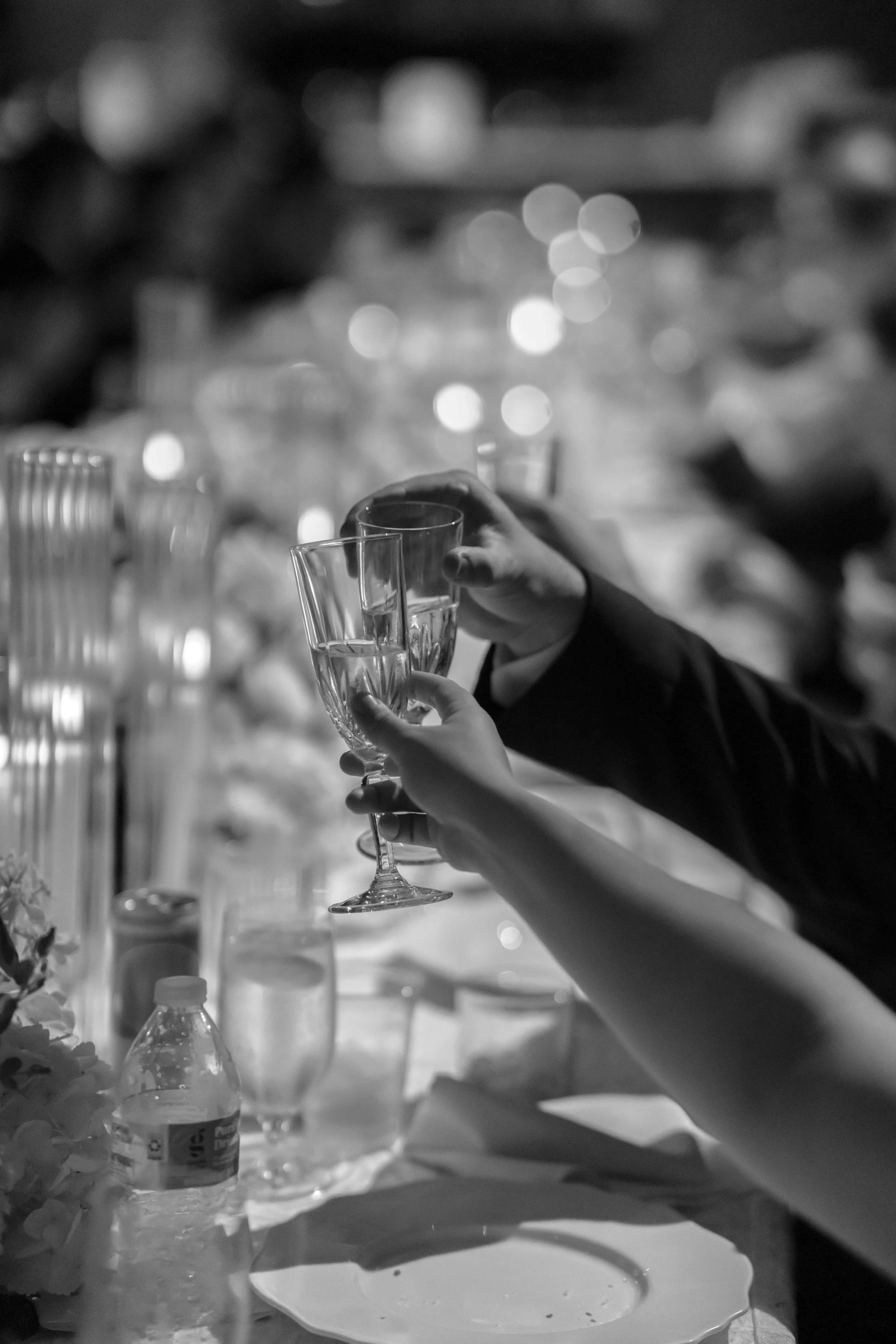 Black and white photo of a person's hand holding a wine glass at a table with various bottles and glasses.