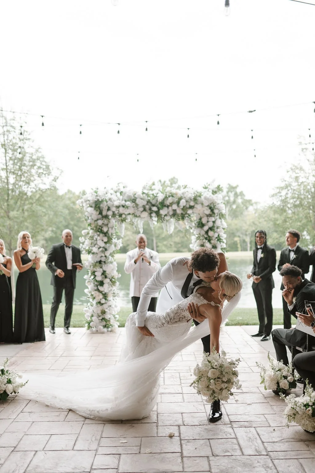 Bride and groom share a romantic dip during their first dance at an elegant indoor wedding ceremony, surrounded by lush white florals and their wedding party.