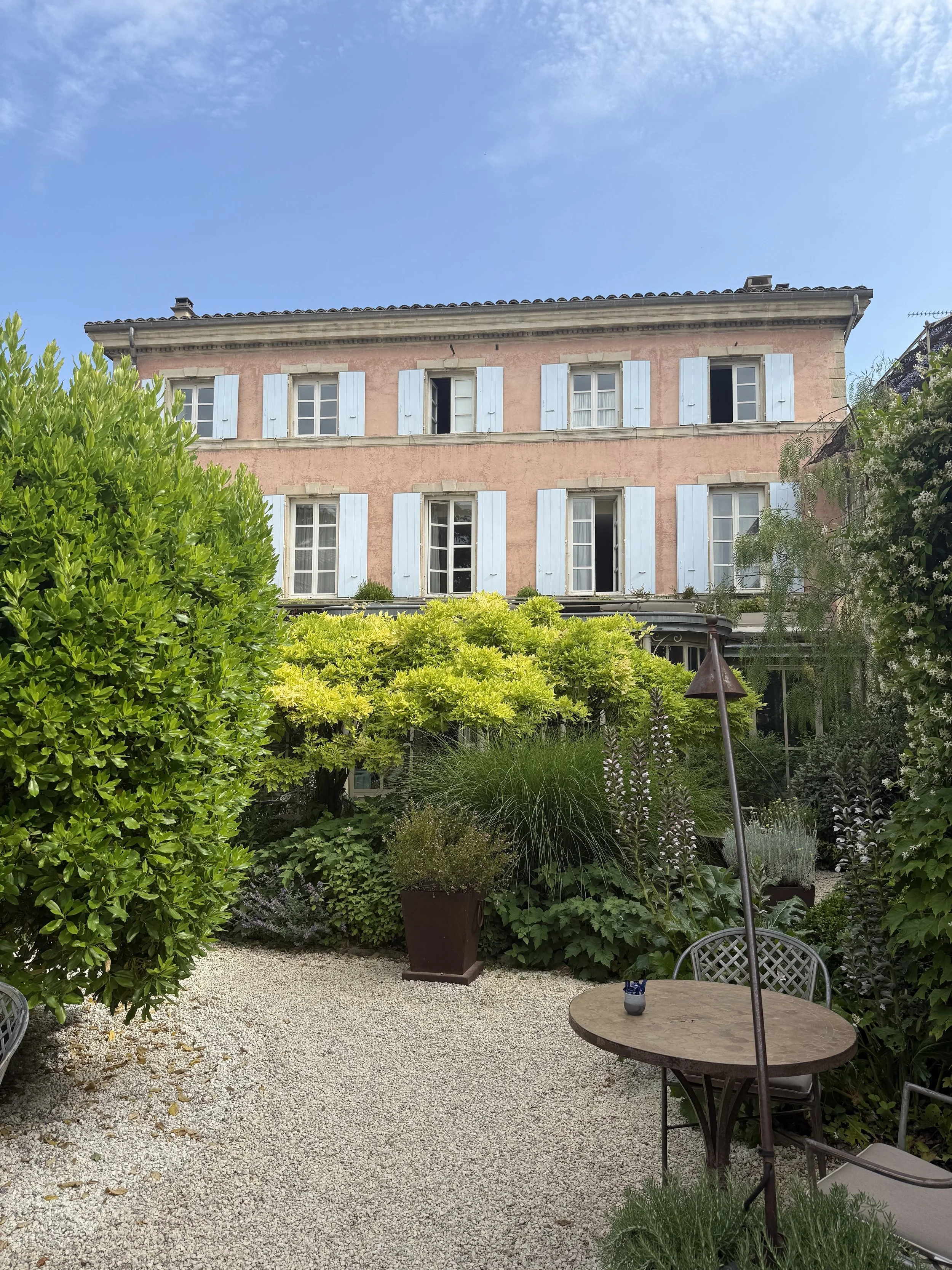 A lush garden patio with a gravel ground, surrounded by various green plants and shrubs, with a pink and white building in the background and a blue sky above.