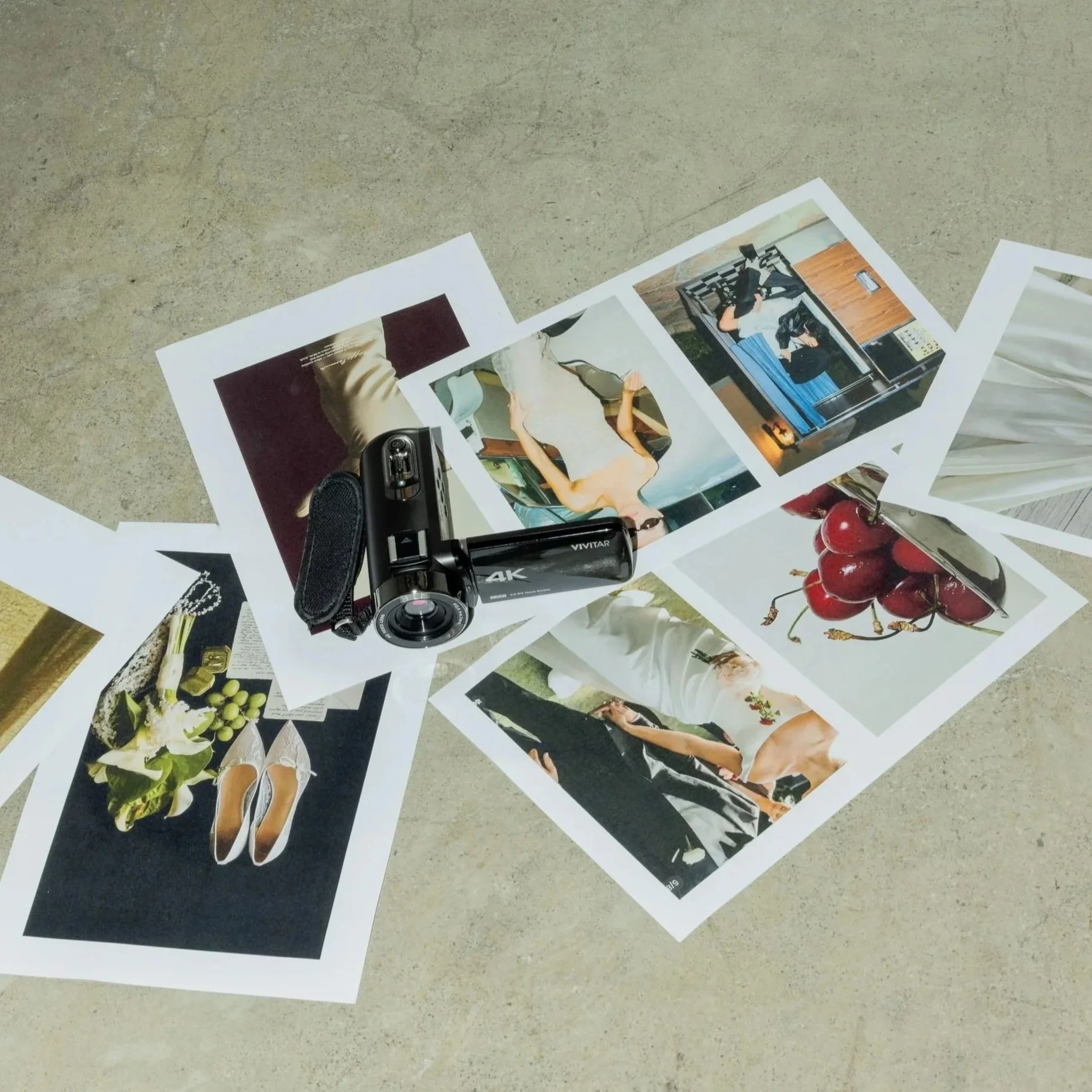 Photo of printed photographs scattered on a surface, including images of people, objects, and scenes, with a video camera and cherries nearby.