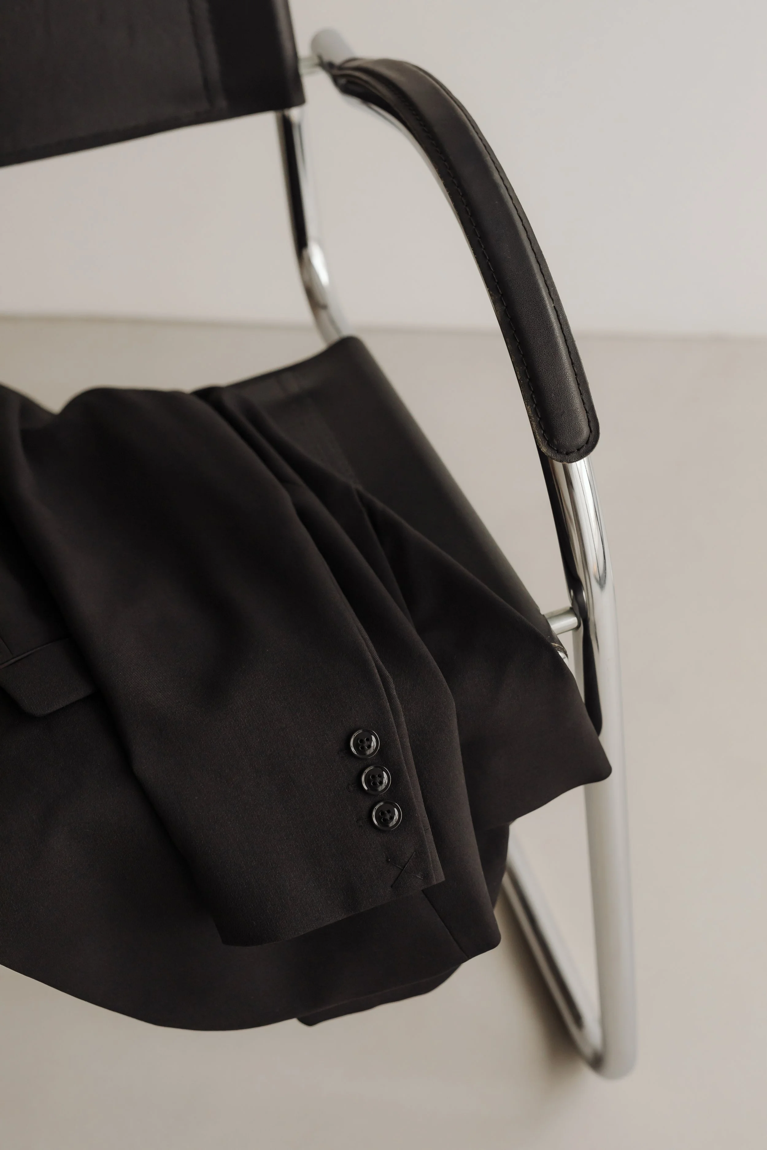 Close-up of a black blazer, displaying buttons and fabric details, hung on a metal and black leather hanger against a plain white background.