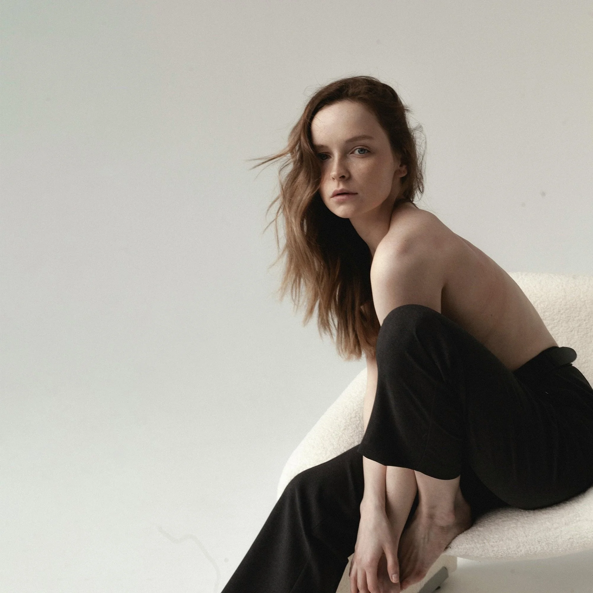 A woman with long brown hair and blue eyes sitting on a white carpeted floor against a beige wall, wearing a black sleeveless dress.