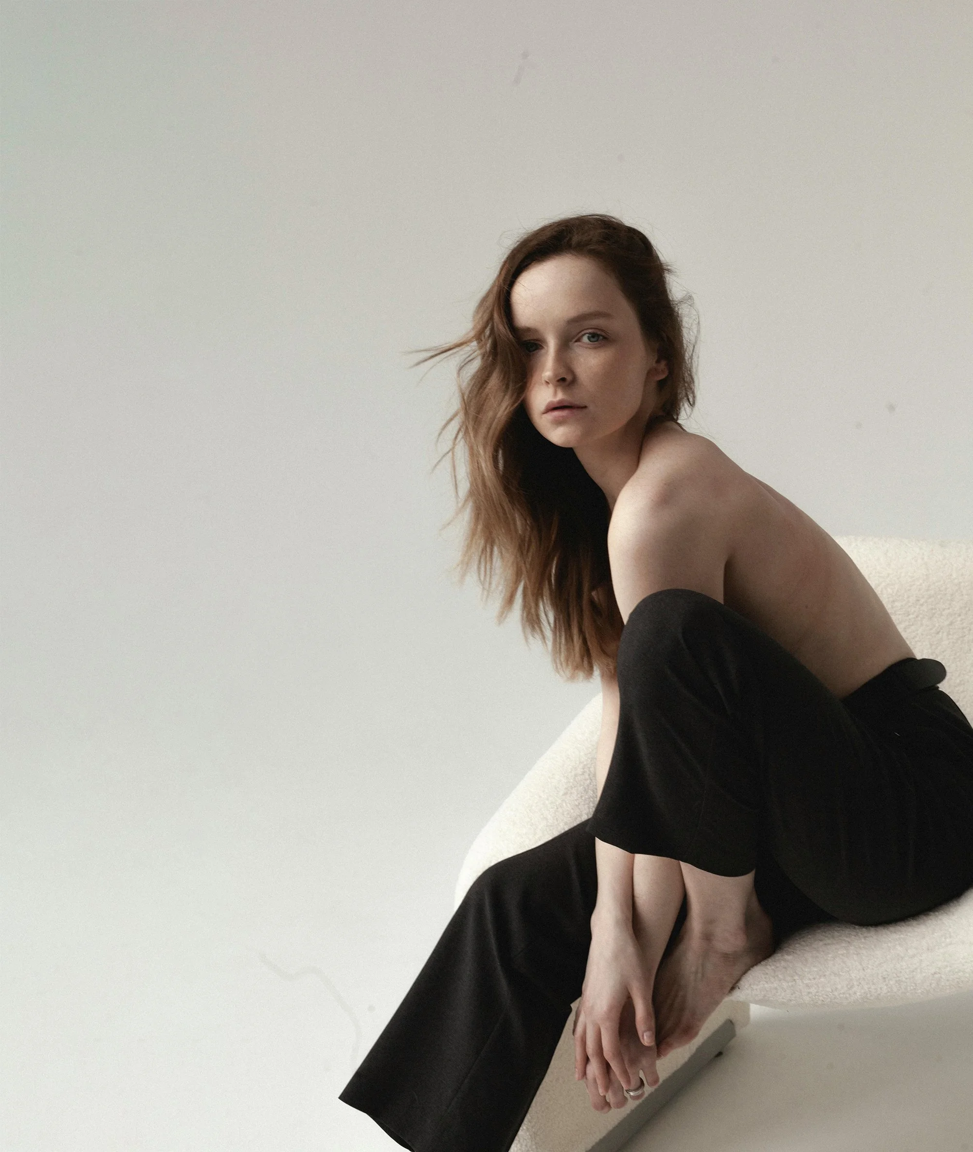 A woman with long red hair sits on a white chair in front of a plain white wall.