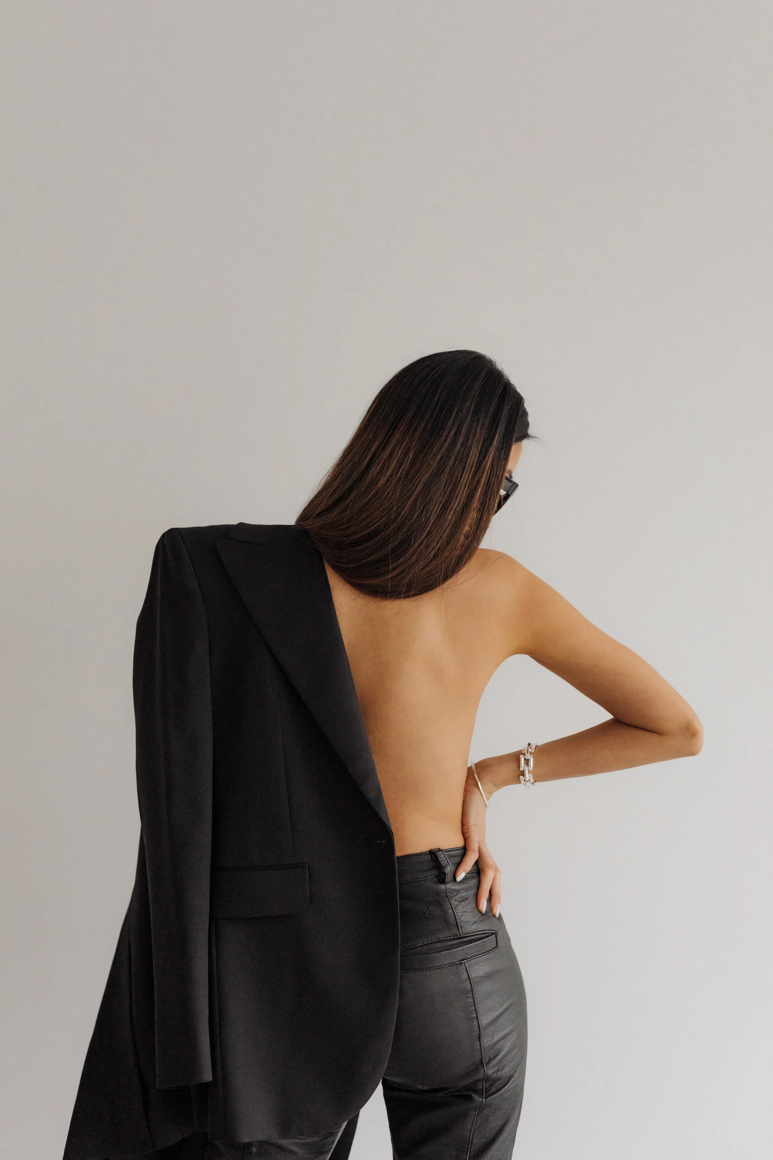 A woman with brown hair and glasses wearing a black blazer drapes over one shoulder, exposing her bare shoulder and arm, while another woman with dark hair and wearing black leather pants touches her waist against a plain gray background.