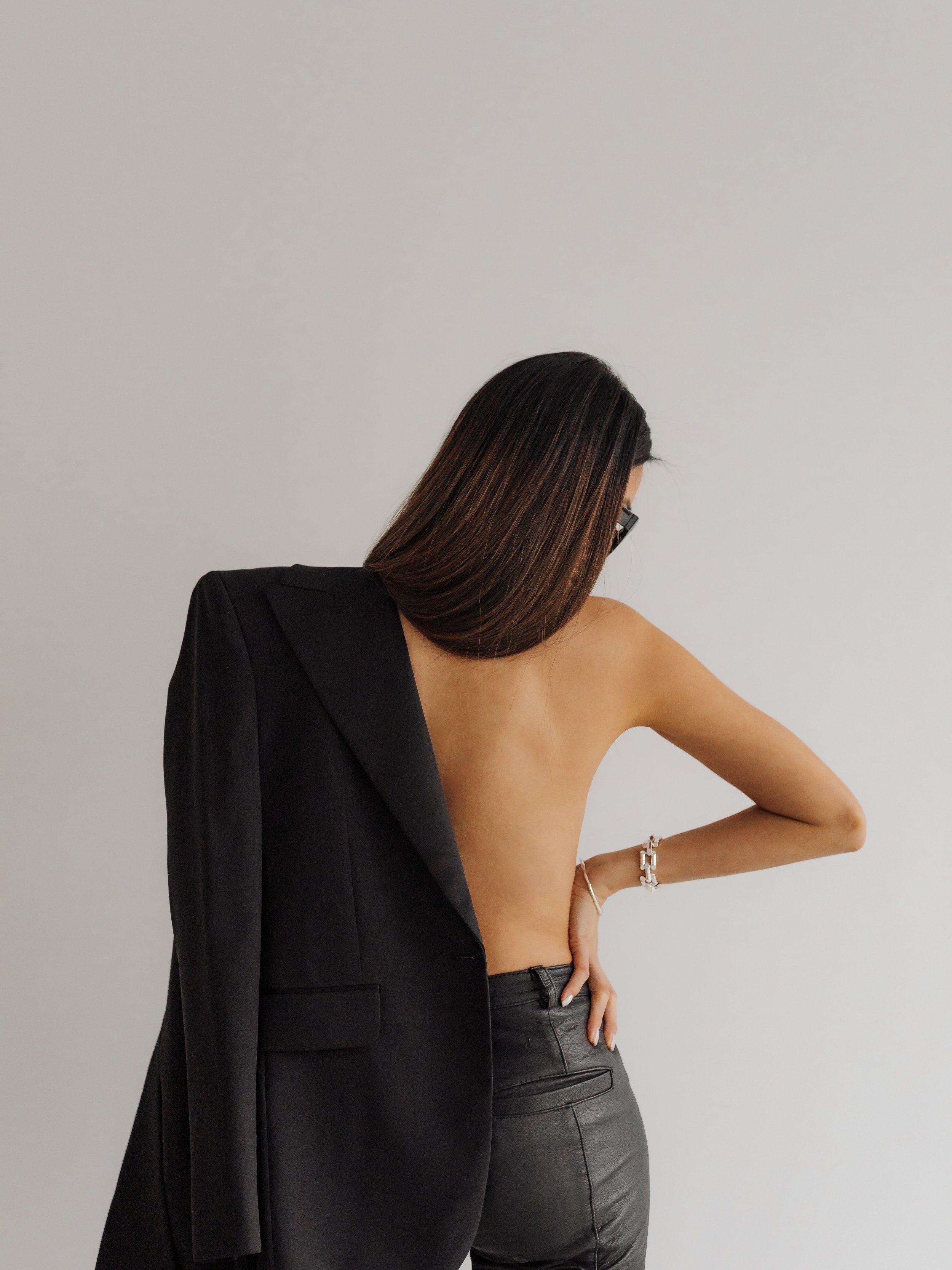 A woman with long dark hair and glasses touching another woman's shoulder. The woman being touched is wearing a black leather skirt and a bracelet, while the woman touching her has a black blazer draped on her shoulder, against a plain light background.