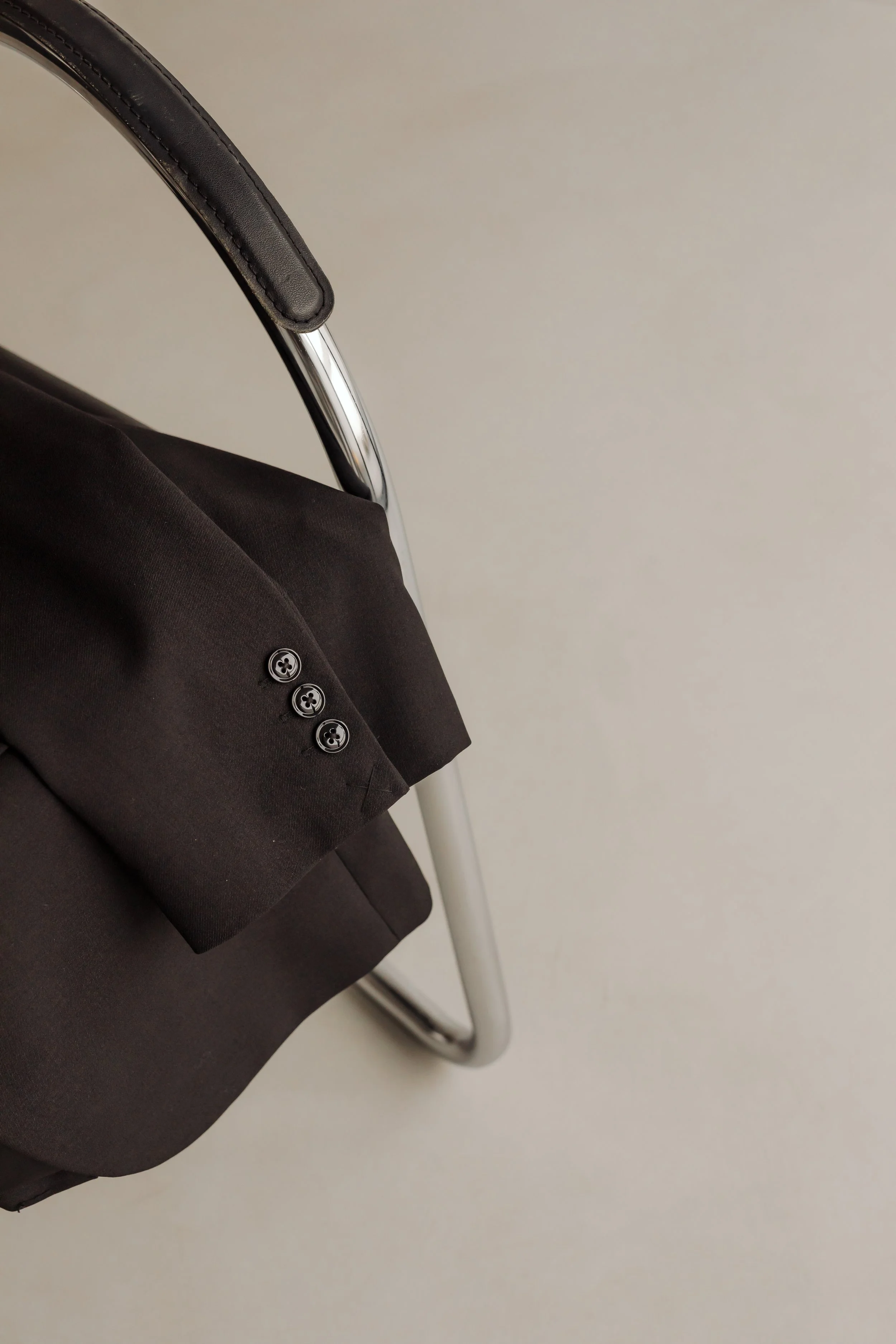 Close-up of a black suit jacket sleeve with three buttons, partially visible, with an armrest of a chair in the background.