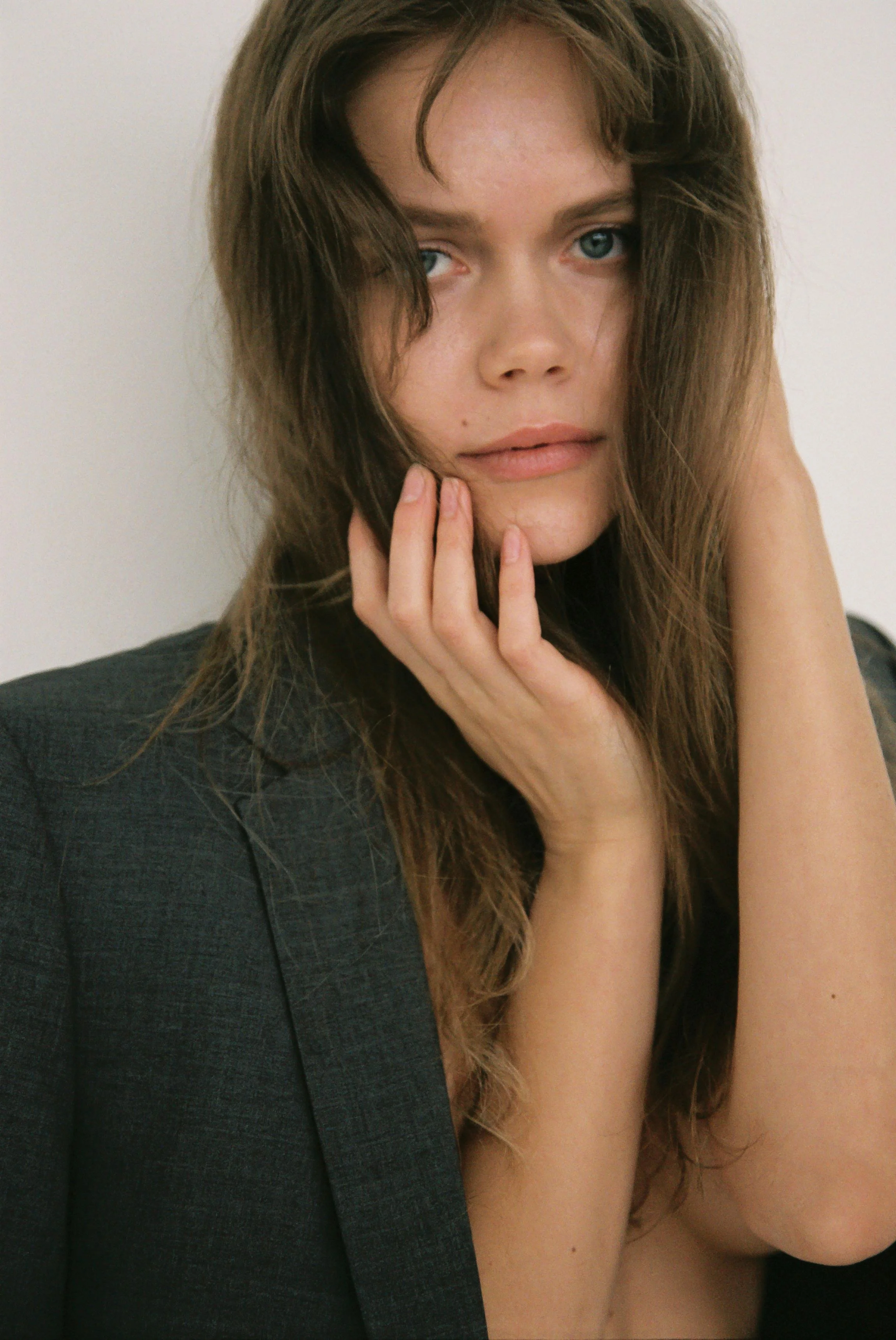A woman with long brown hair and light skin, partially topless, resting her hand on her chin and cheek, looking directly at the camera with a serious expression, against a plain white background.
