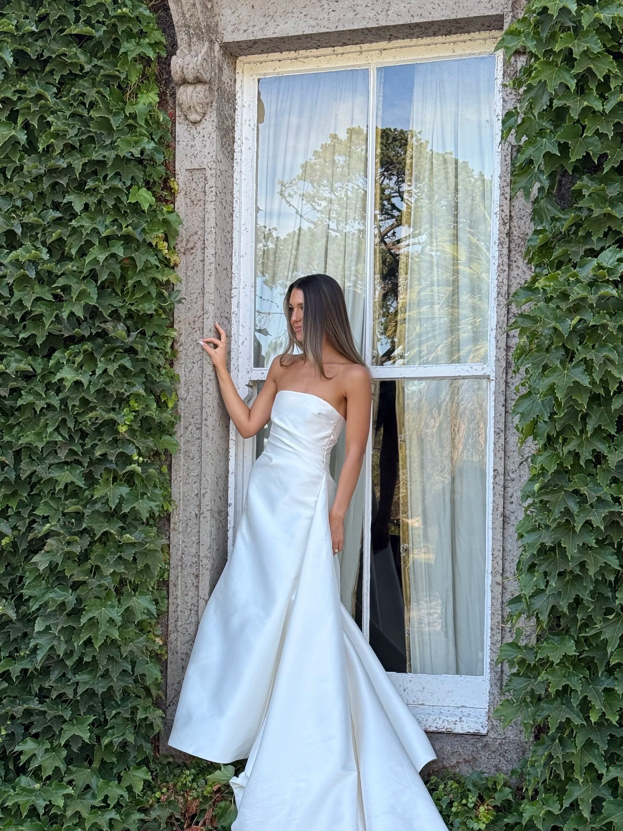 A woman in a white strapless gown standing in front of a window surrounded by green ivy.