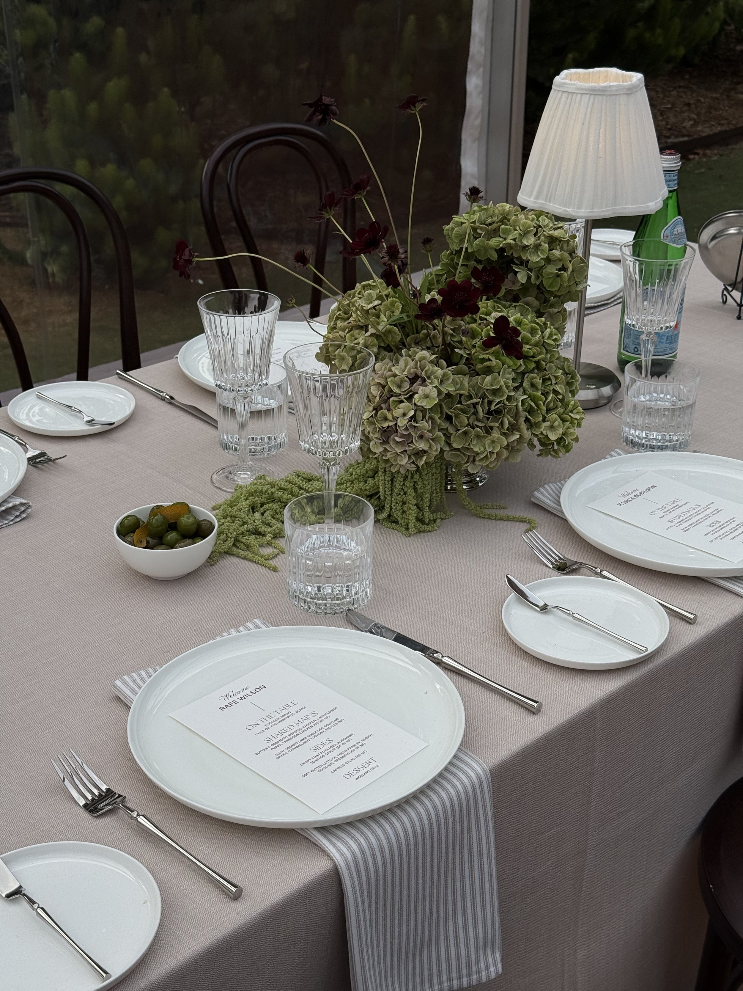 A table set for a meal with white plates, silverware, glassware, a bowl of green and dark-colored olives, and a floral centerpiece with green and dark red flowers. There is a small white table lamp, a green glass bottle, and a printed menu on the plates. The tablecloth is a light beige color, and the chairs are black with wooden backs.