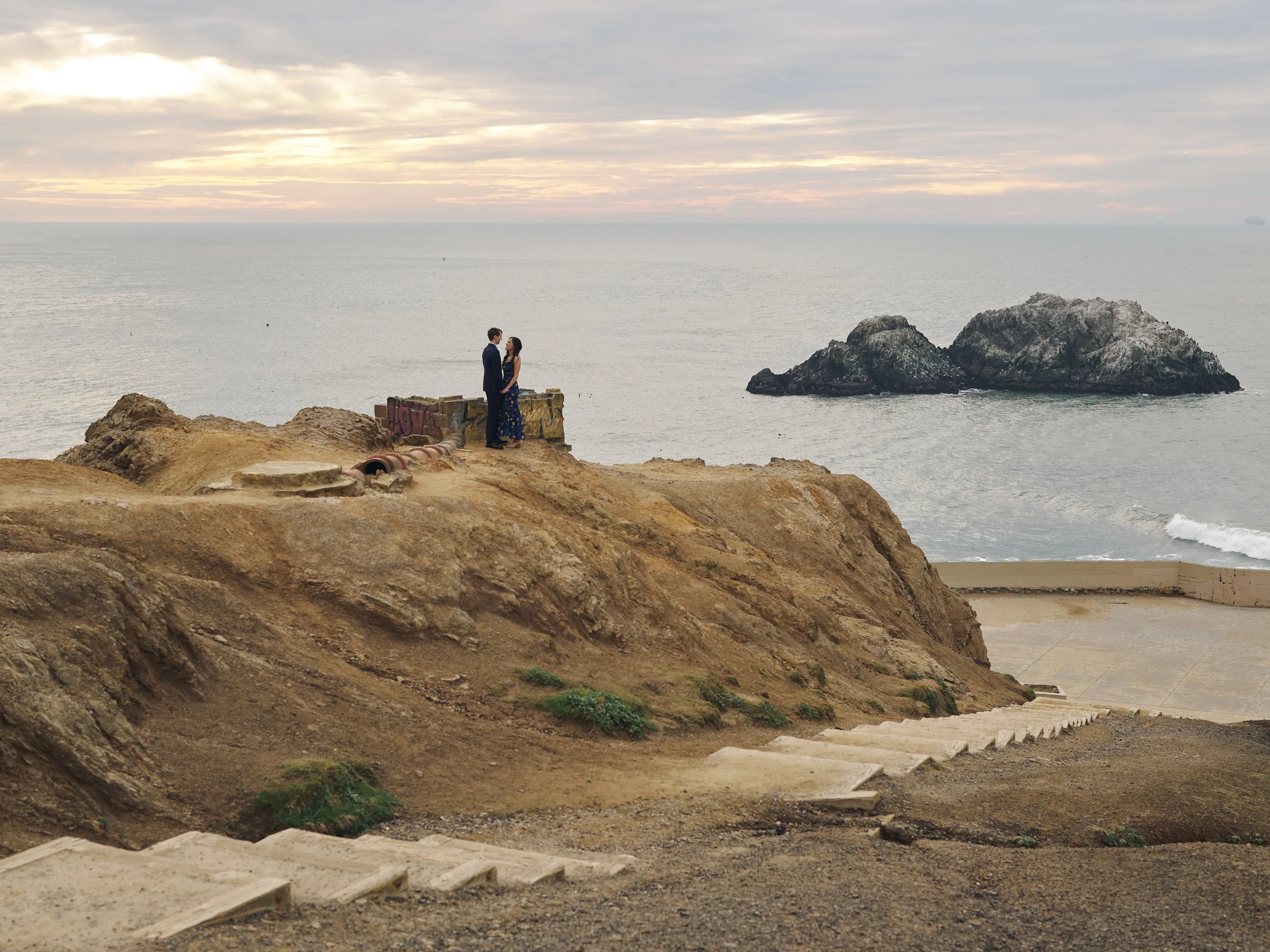 Couple small in the wide landscape of Sutro Baths, the Pacific stretching behind them