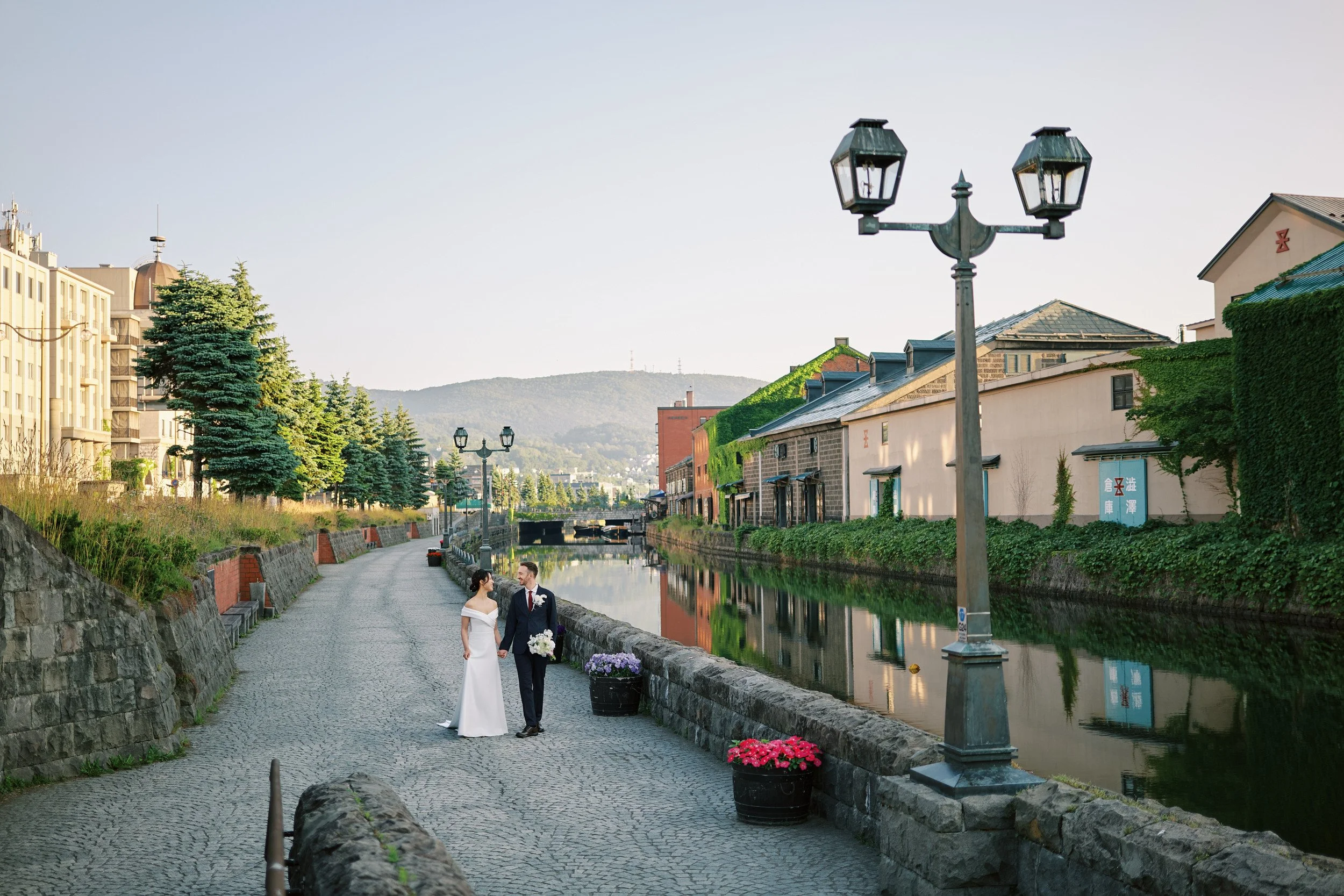 Couple standing together along the Otaru Canal at sunrise, stone walkway and historic warehouses reflected in still water
