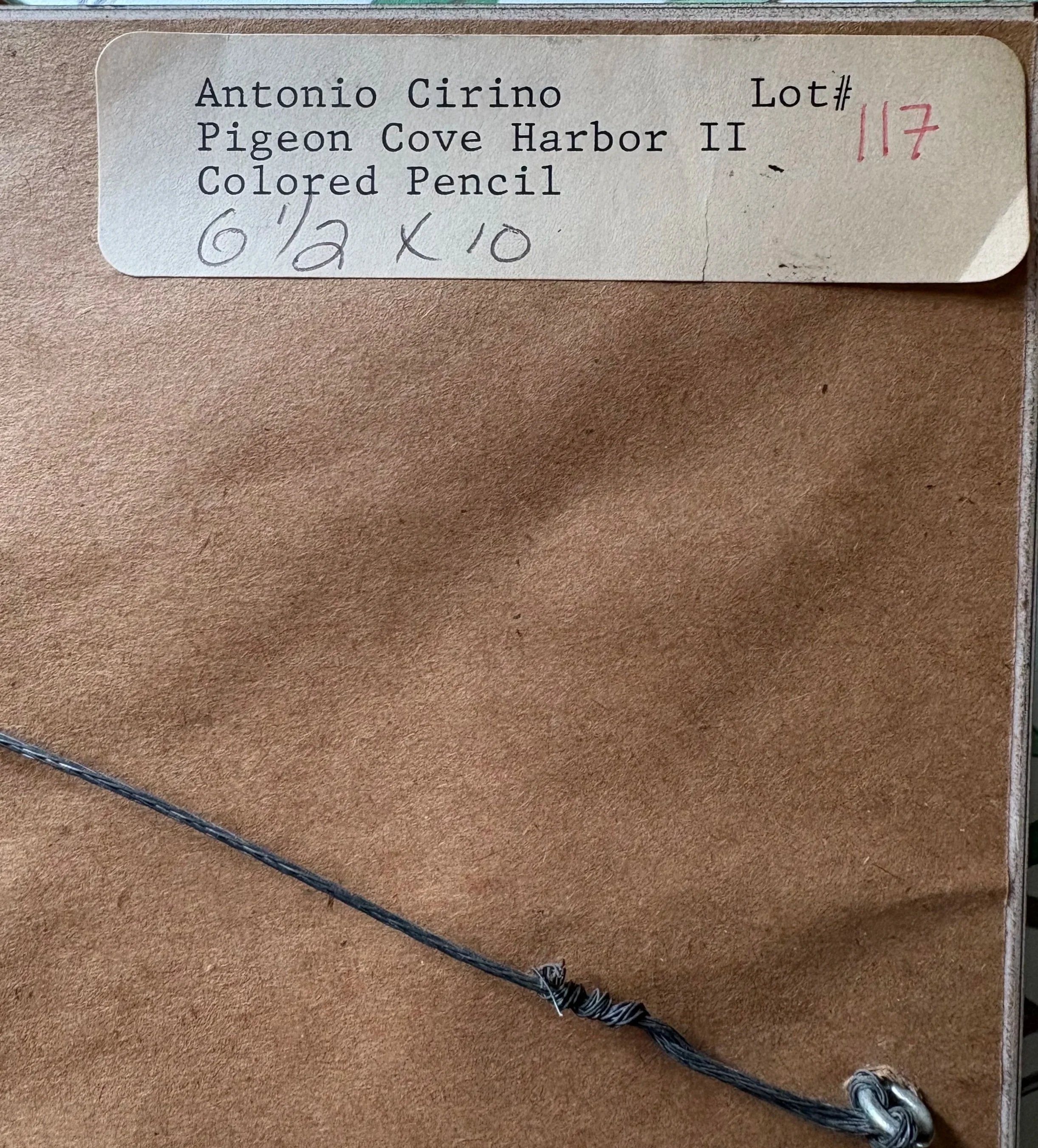 A label with typed and handwritten text for a fishing lure, reading 'Antonio Cirino, Lot# 117, Pigeon Cove Harbor II, Colored Pencil, 6 1/2 X 10', attached to a fishing lure on a wooden surface.