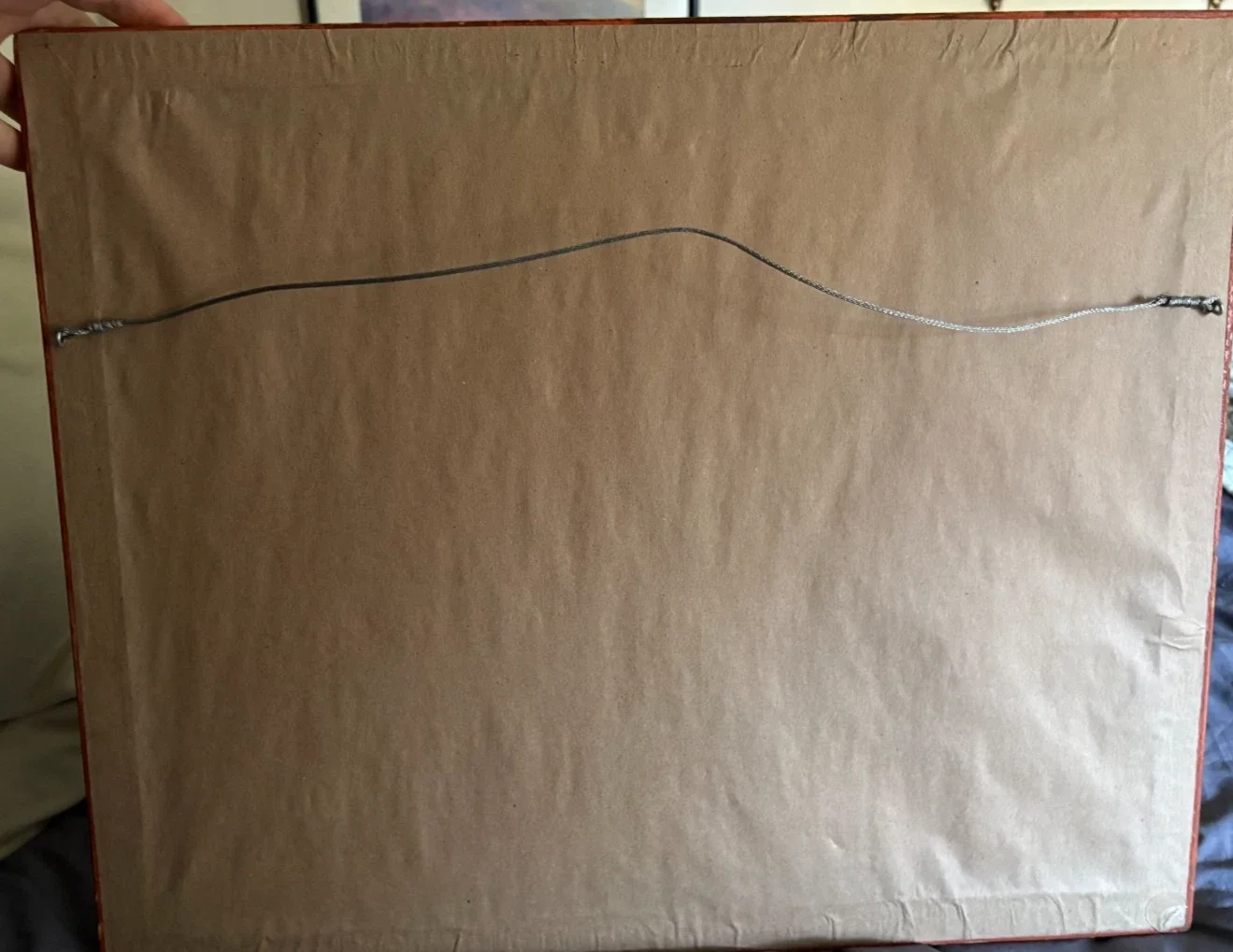 Back of a framed item with a visible wire for hanging, attached to two hooks on each side of the frame.