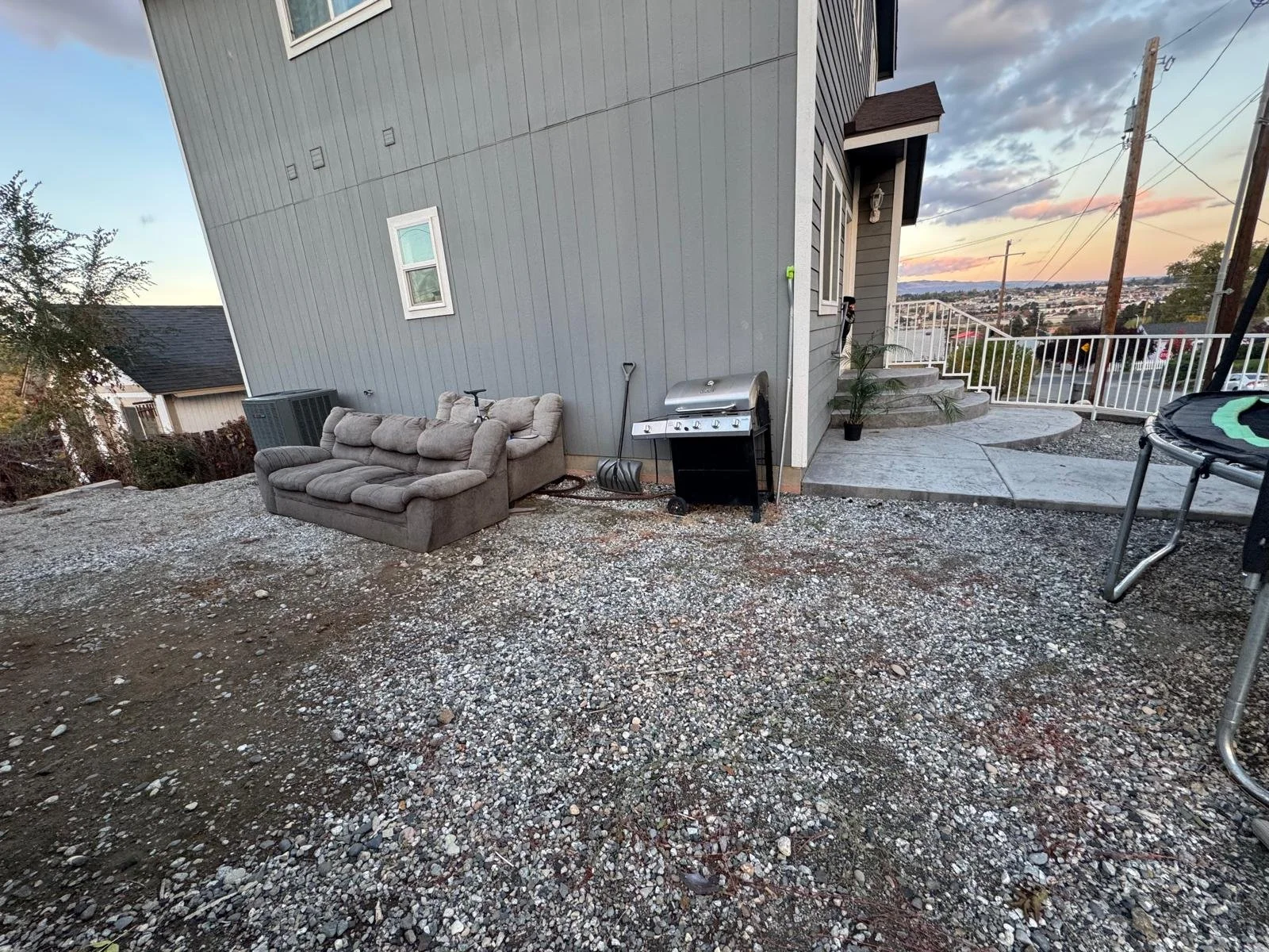 Backyard patio area with a gray couch, a grill, a snow shovel, and a trampoline, with a house and a view of the city in the background during sunset.