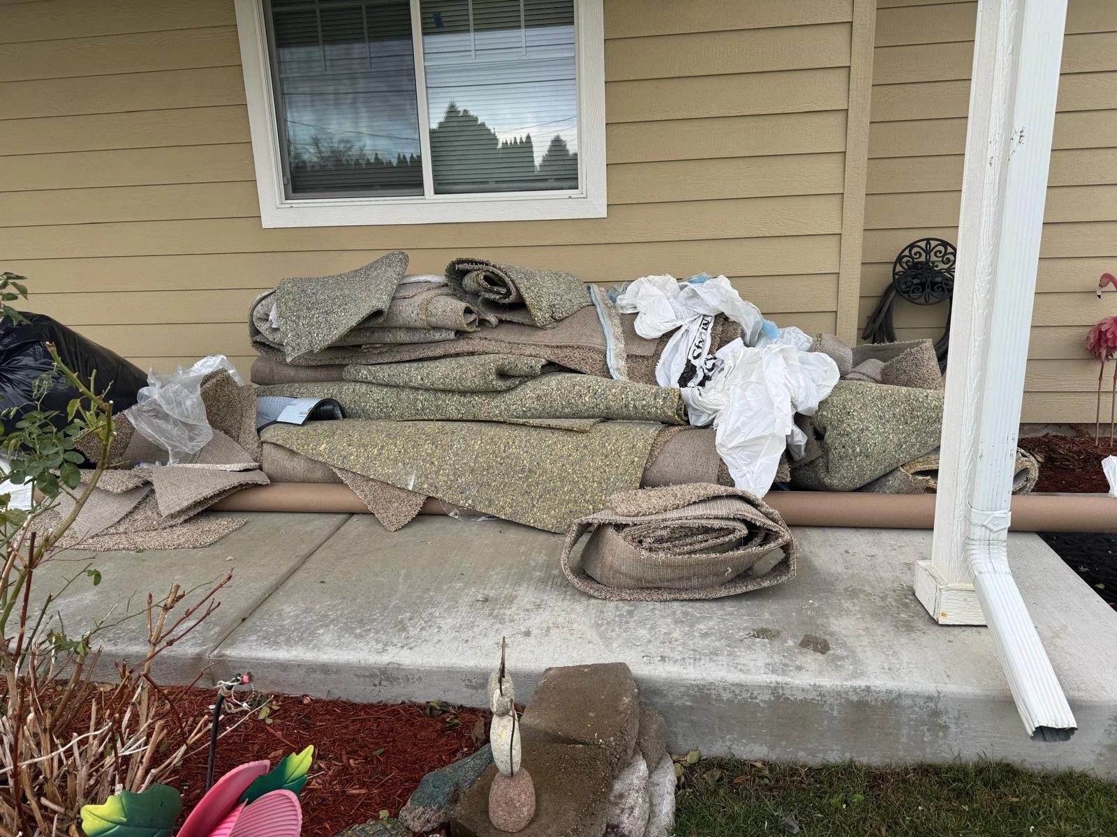 Piles of rolled-up and unfolded carpets and rugs on a concrete porch next to a house wall. Some plastic bags and a black item are also visible among the carpets.