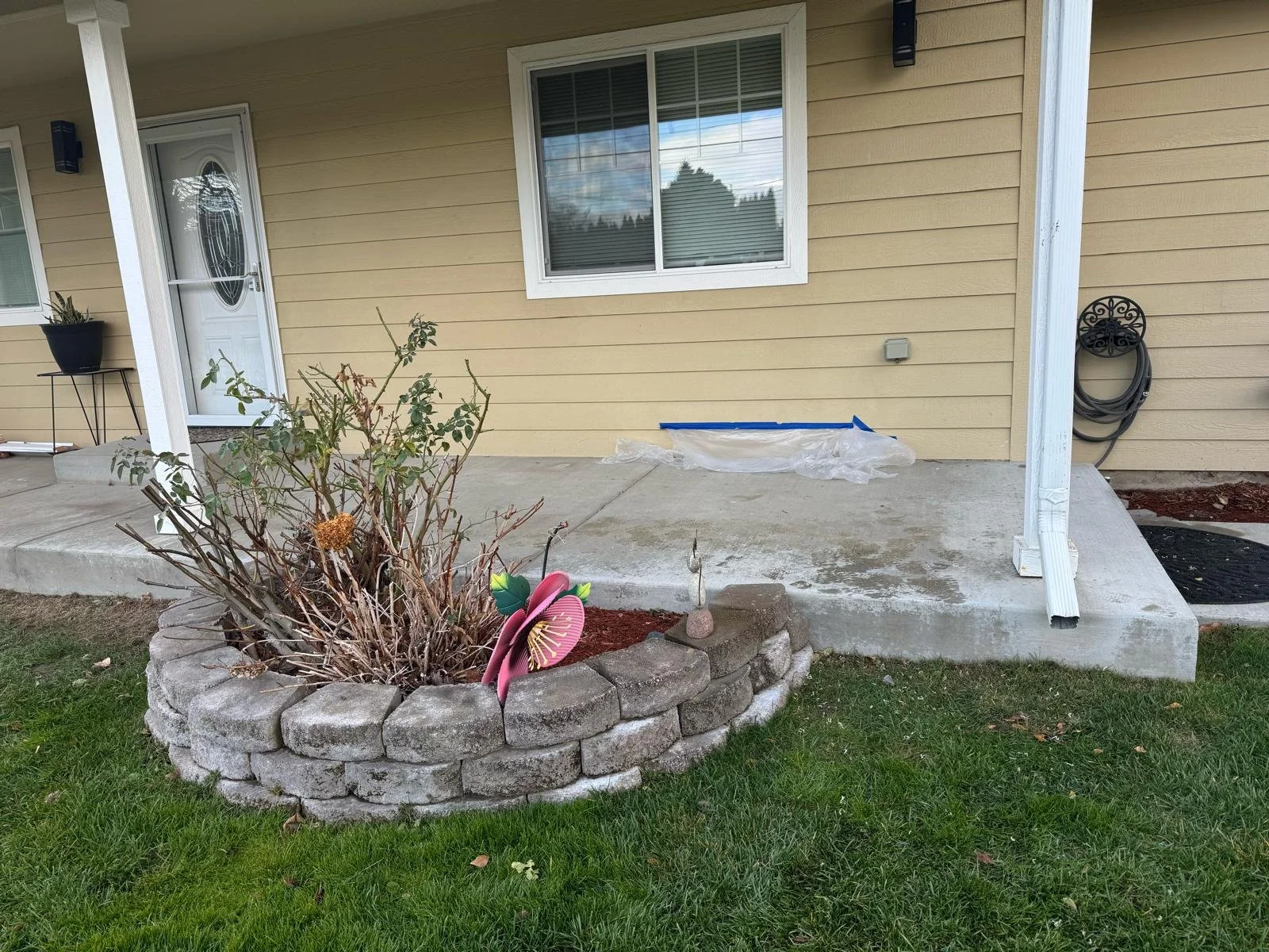 Back porch with beige siding, a window, white door, potted plant, and a black hose reel. There is a small garden bed with a bush and decorative pink flower in a stone border, and a concrete step leading to the porch.