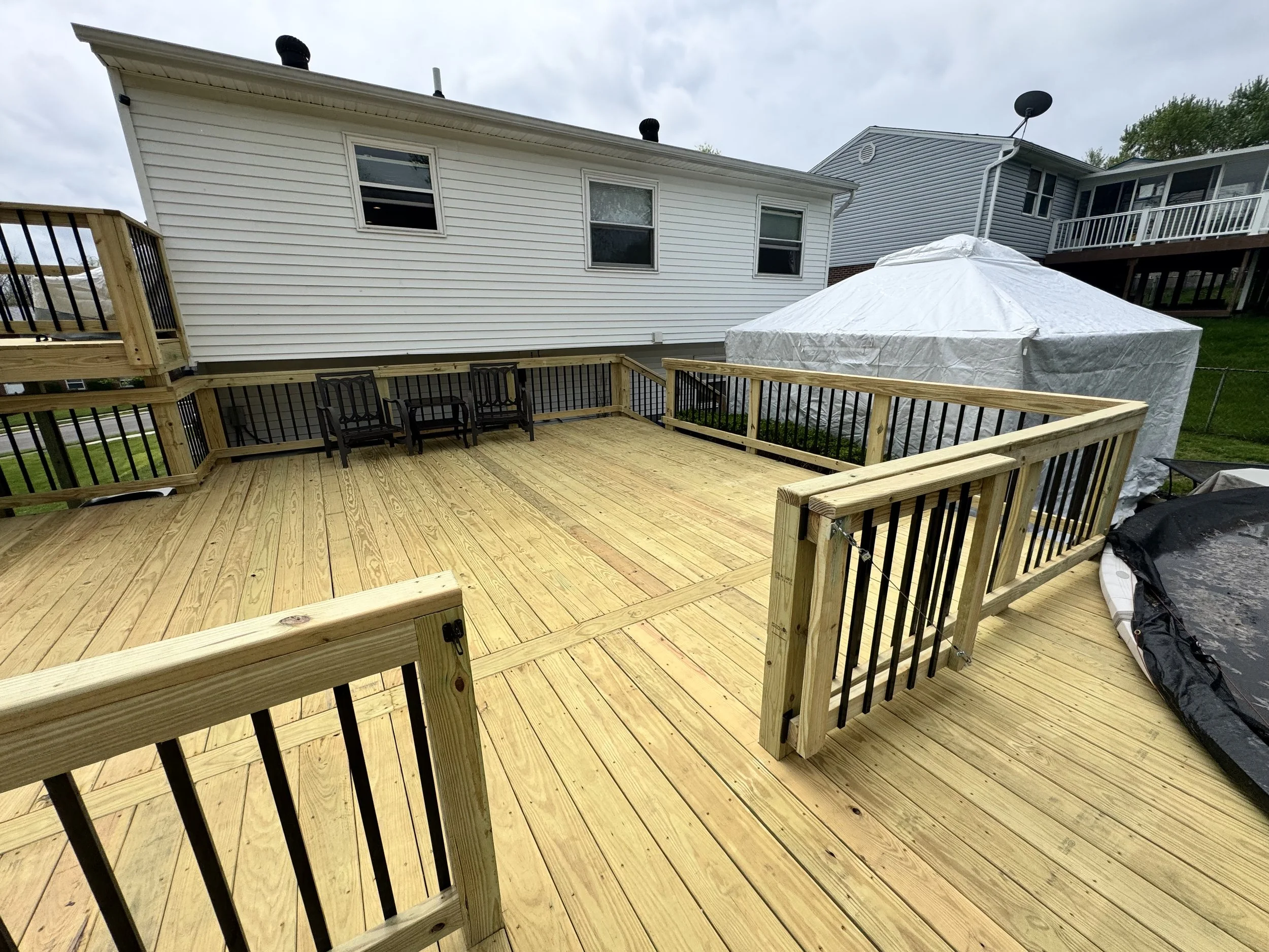 A backyard deck with unfinished wooden flooring, black metal railings, a few black chairs, and a white covered object, possibly a hot tub. The house has white siding, three windows, and a neighboring house with a balcony and a satellite dish.