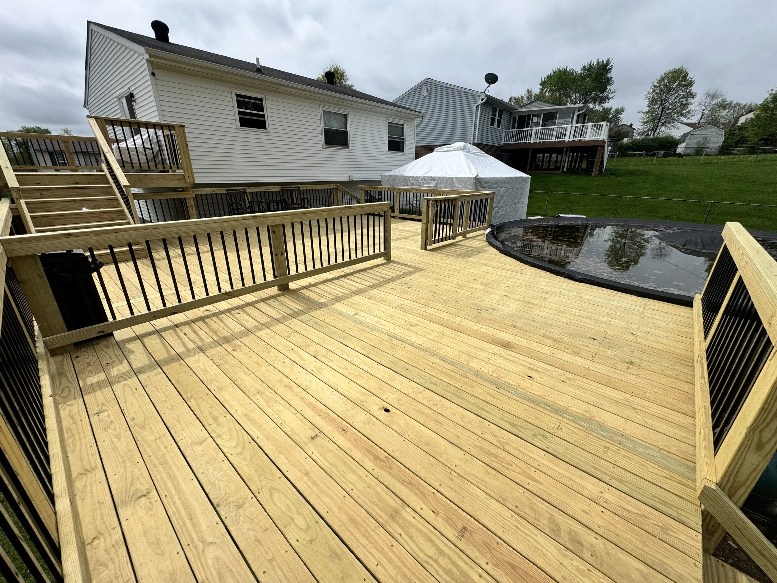 A backyard deck with newly installed wooden flooring, surrounded by a black metal railing. There's a small pool covered with a black pool cover, and a white patio umbrella nearby. In the background, there are neighboring houses, trees, and a grassy yard under an overcast sky.