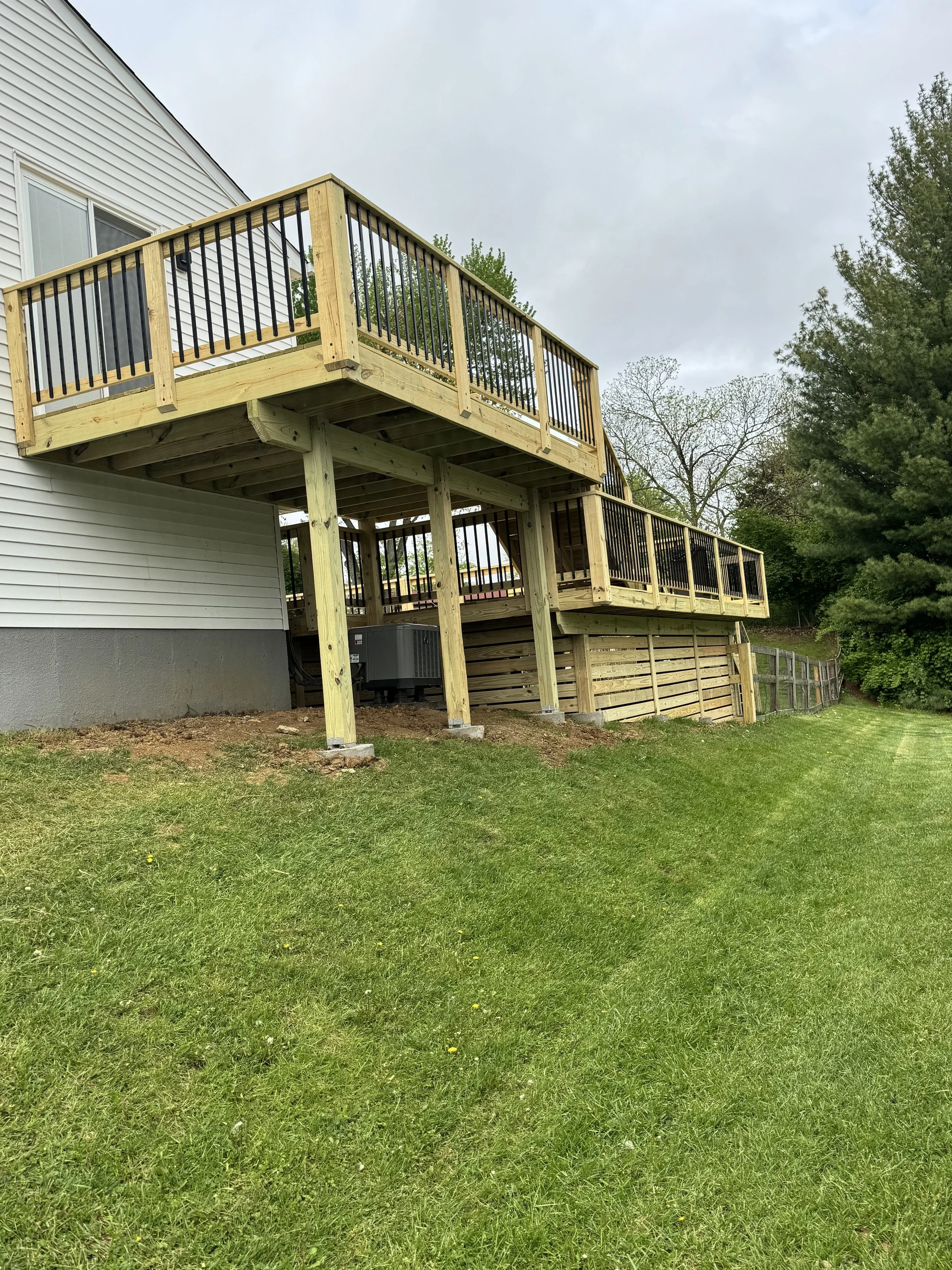 A newly built wooden deck attached to a white house, with black metal railings, overlooking a green grassy yard with trees in the background.