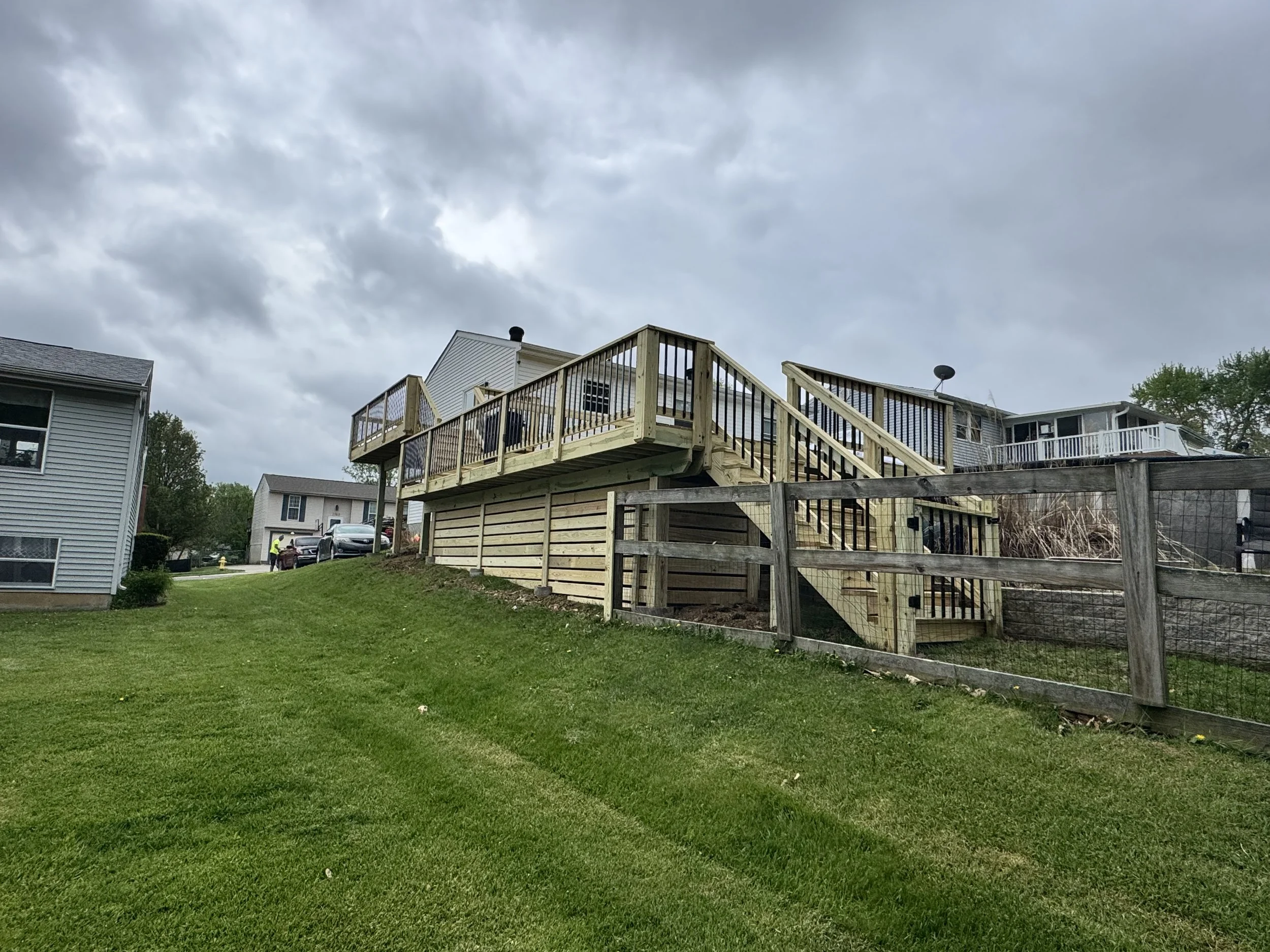 A residential backyard with a newly built wooden deck attached to a two-story house. The yard has green grass and a wooden fence on the right side. Overcast skies with dark clouds are seen in the background, and there are neighboring houses visible in the distance.