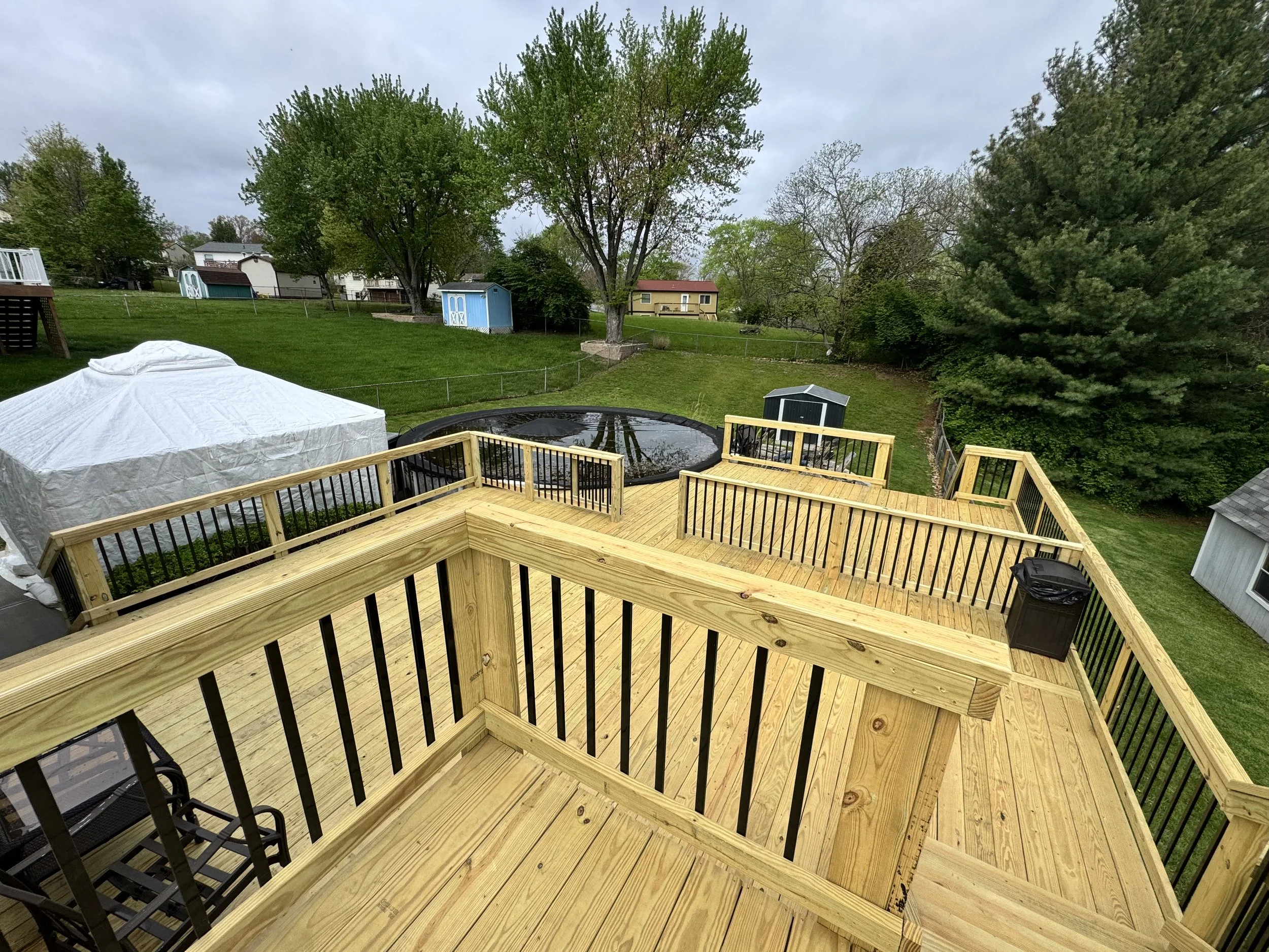 A backyard deck with a railing overlooking a lawn with trees, small sheds, and a trampoline.