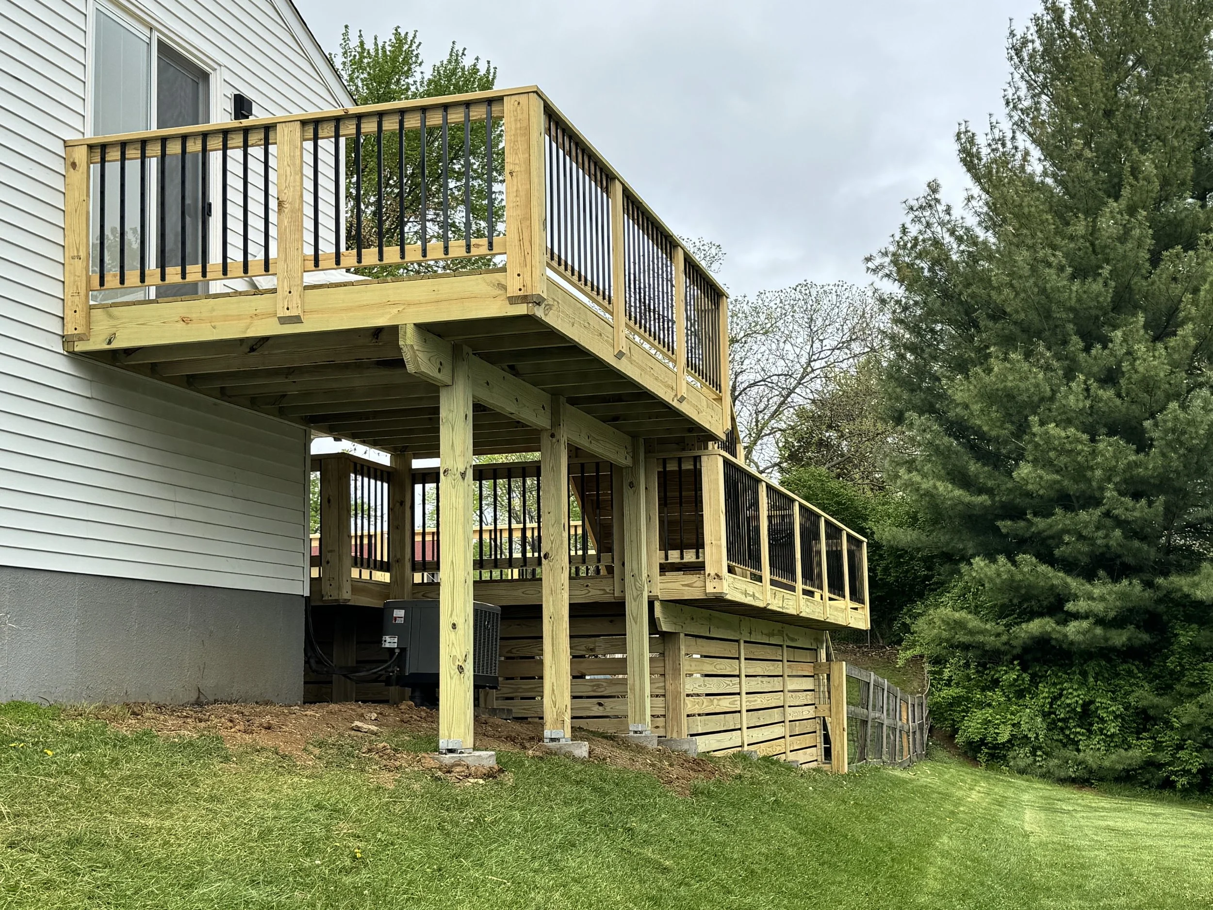 A two-story house with white siding has a new wooden deck with black metal railings attached to the back. The deck is supported by wooden posts and has stairs leading down to a grass yard. Surrounding trees are visible in the background.