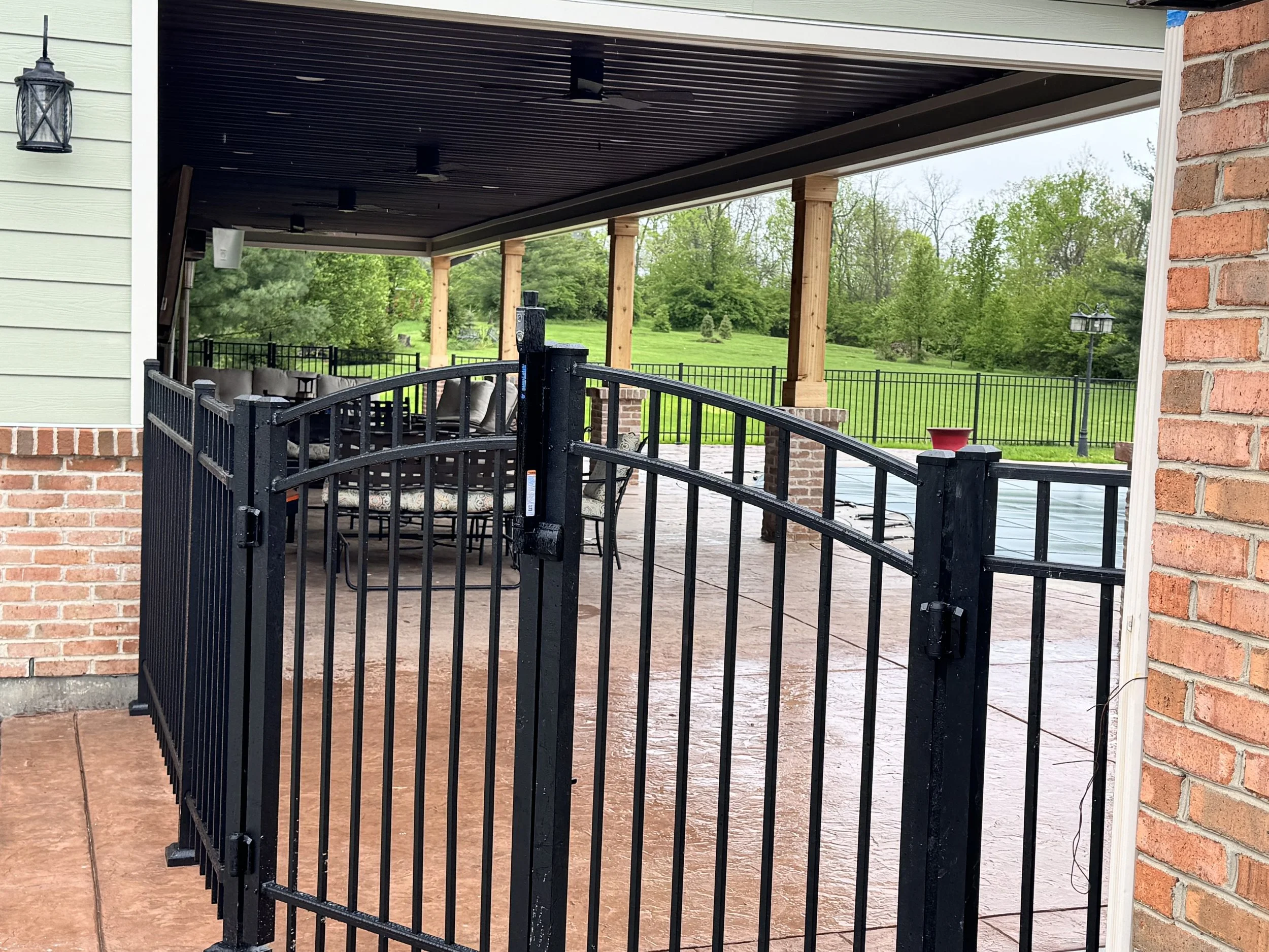 A covered outdoor patio area with a black metal fence, patio furniture, and a view of green trees and grass.