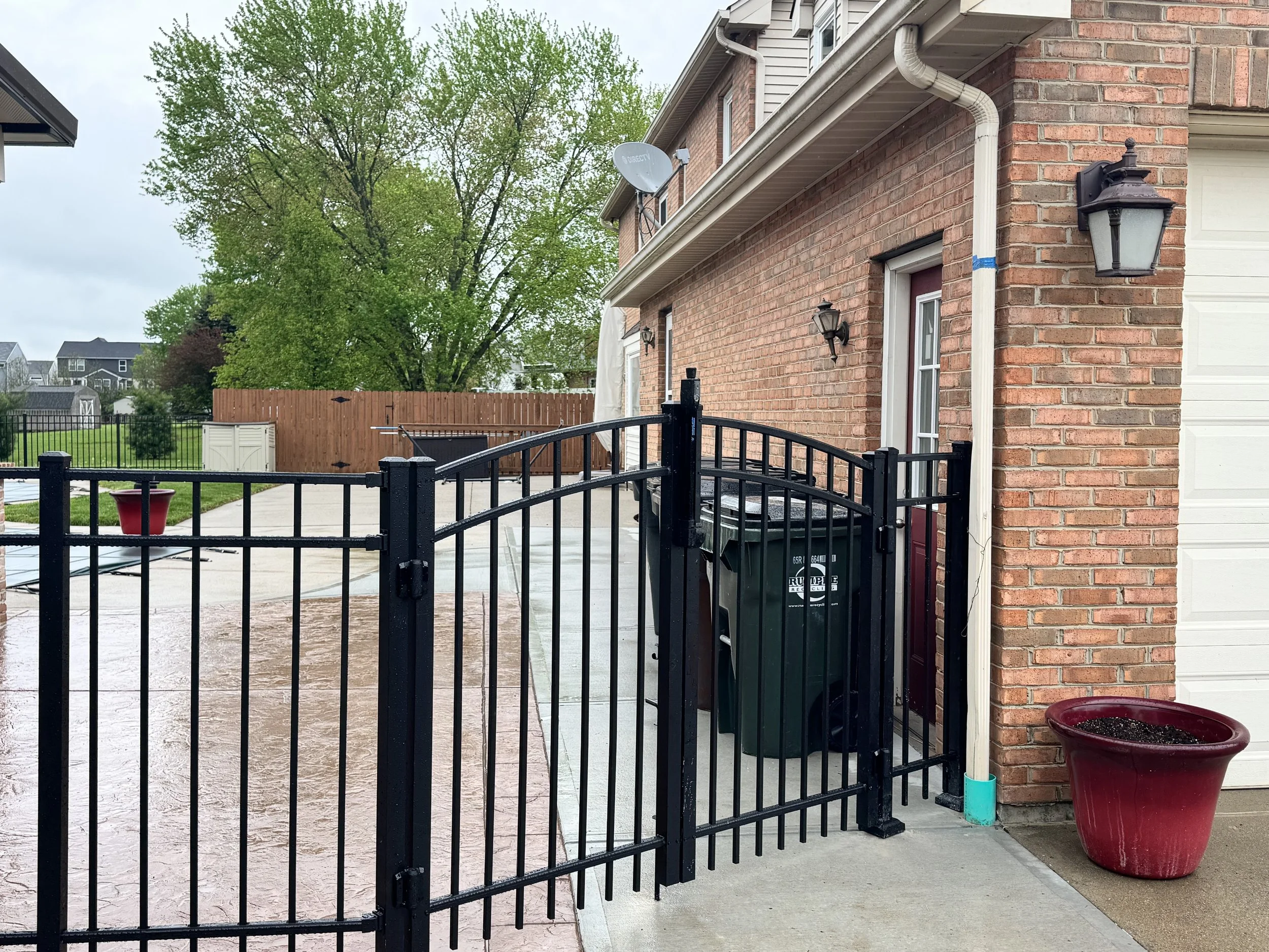 View of a backyard patio area with a black metal gate, a large red flowerpot, a fence, green trees, and a brick house exterior with a satellite dish and outdoor lights.