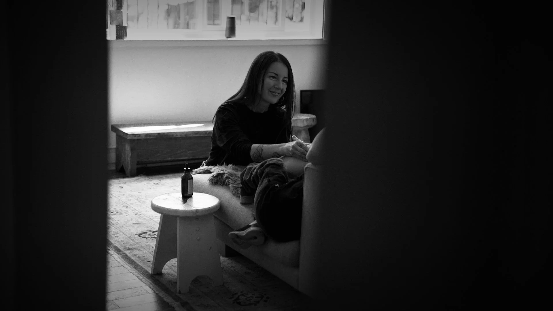 A woman smiling and sitting on a sofa during a therapy session, with a therapist's legs visible, a small round table with a bottle on it, a window with a bottle, and a rug in the background.
