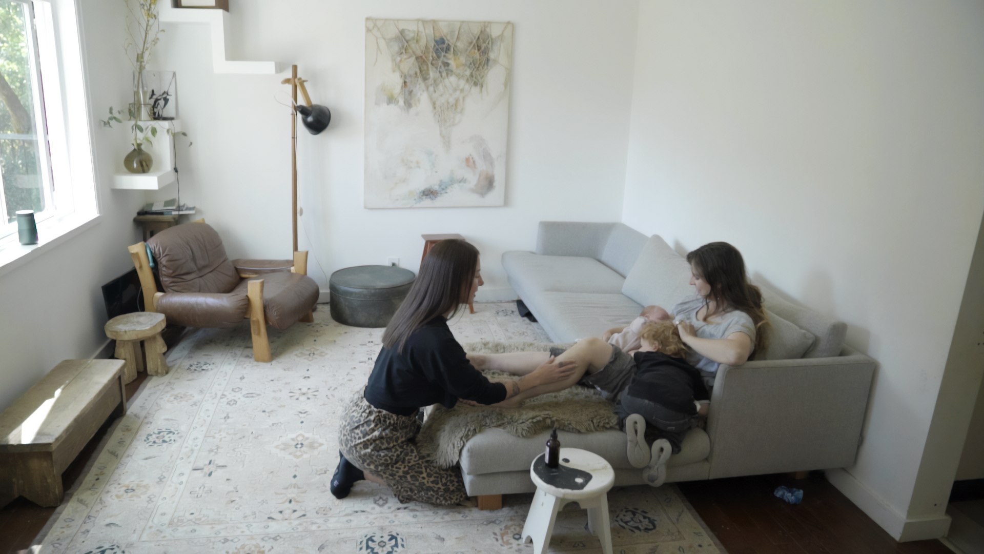 Two women,Tania Marie and a new mom. The new mom sitting on a sofa and Tania Marie is kneeling in front of her preforming her In-Home Postpartum care to her clients legs, with children, in a living room.