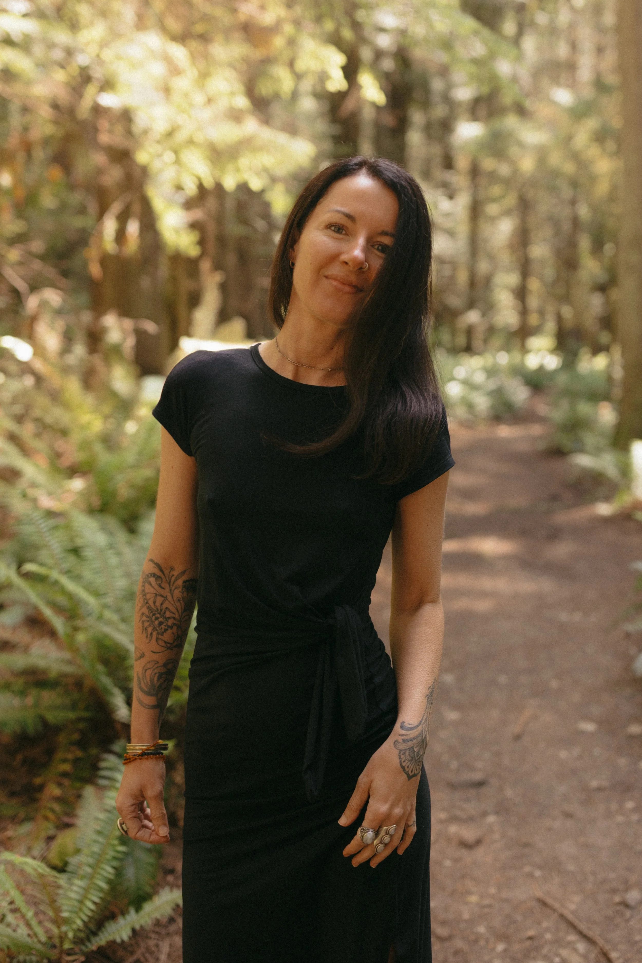 Tania Marie standing on a forest trail with trees and ferns in the background. Tania Edwards, Intuitive guide, supports women through big life transitions with bodywork, ritual, lifestyle guidance, herbal support, and compassionate presence.
