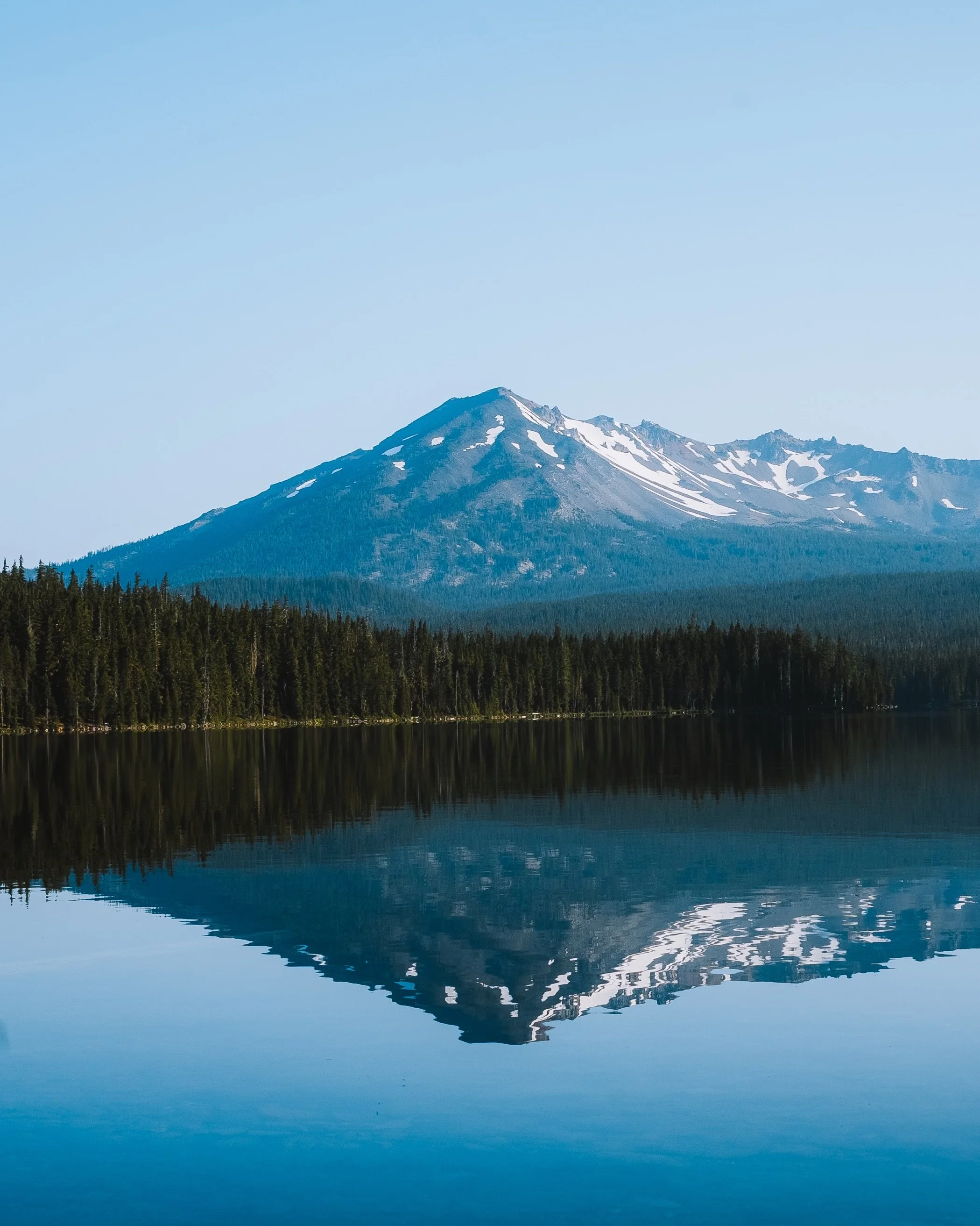 Snow-capped mountain reflected in a calm lake surrounded by a forest.