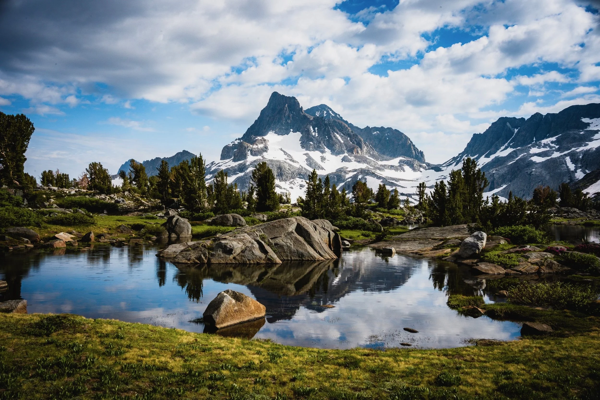 Snow-capped mountain peaks under a partly cloudy sky, with a foreground of green grass, trees, rocks, and a calm reflective lake.