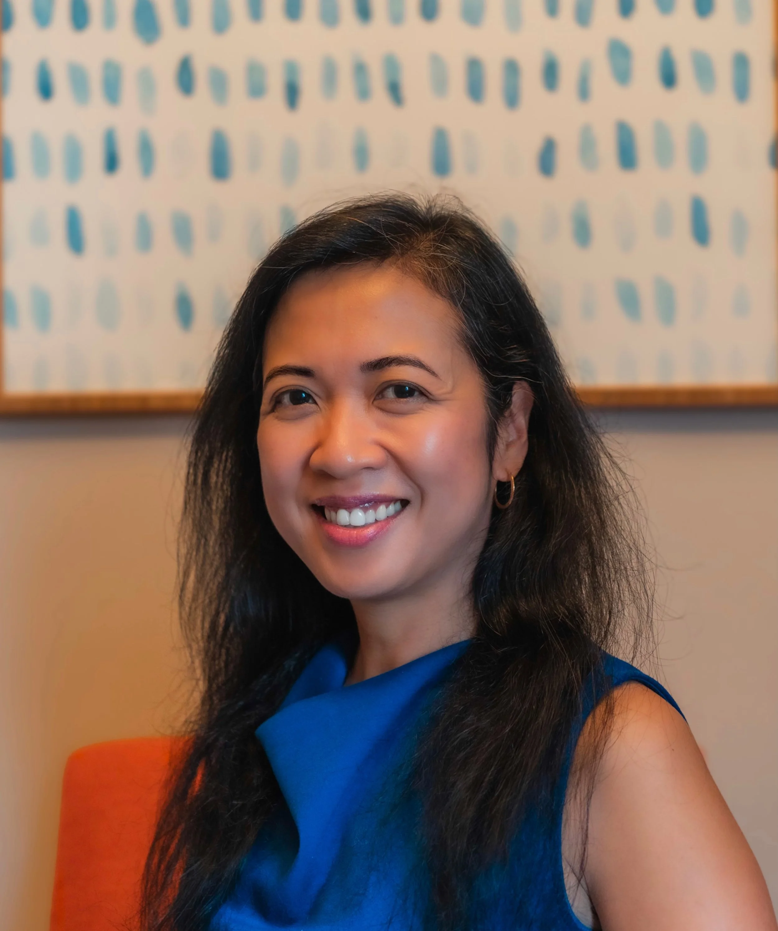 Close-up of a smiling woman with long dark hair sitting against an abstract blue and white patterned wall.