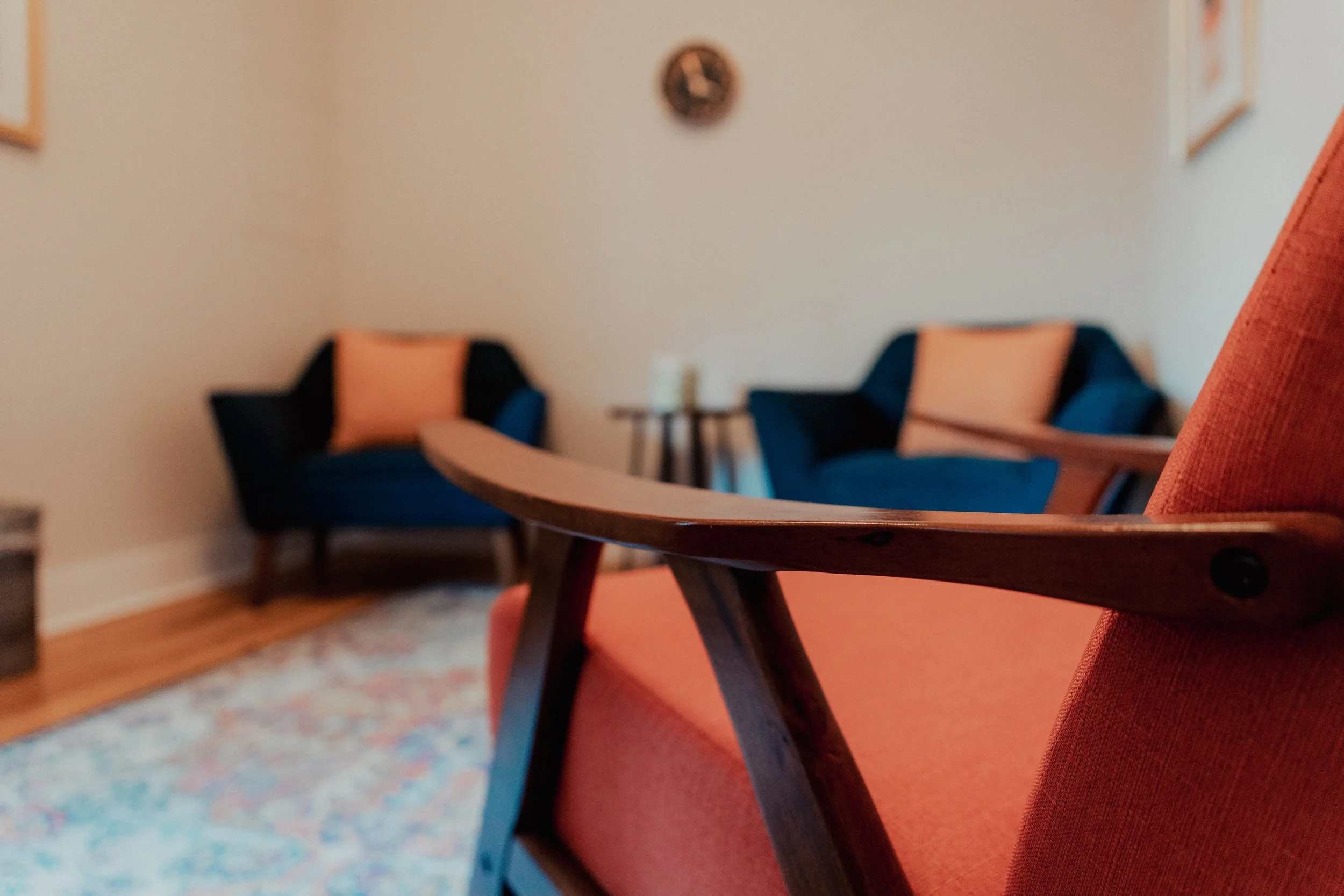 Close-up of a wooden chair with dark wood legs and a burnt orange fabric seat and backrest. In the background, two armchairs with blue fabric and pink cushions are visible, along with a small side table holding a lamp. The room has a softly lit wall with a round clock and framed pictures.