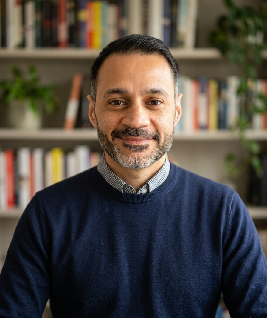 A middle-aged man with short black hair, a beard, and wearing a dark blue sweater over a collared shirt, sitting indoors in front of a bookshelf filled with colorful books and some green plants. Expert clinician knowledgable kind psychiatrist care