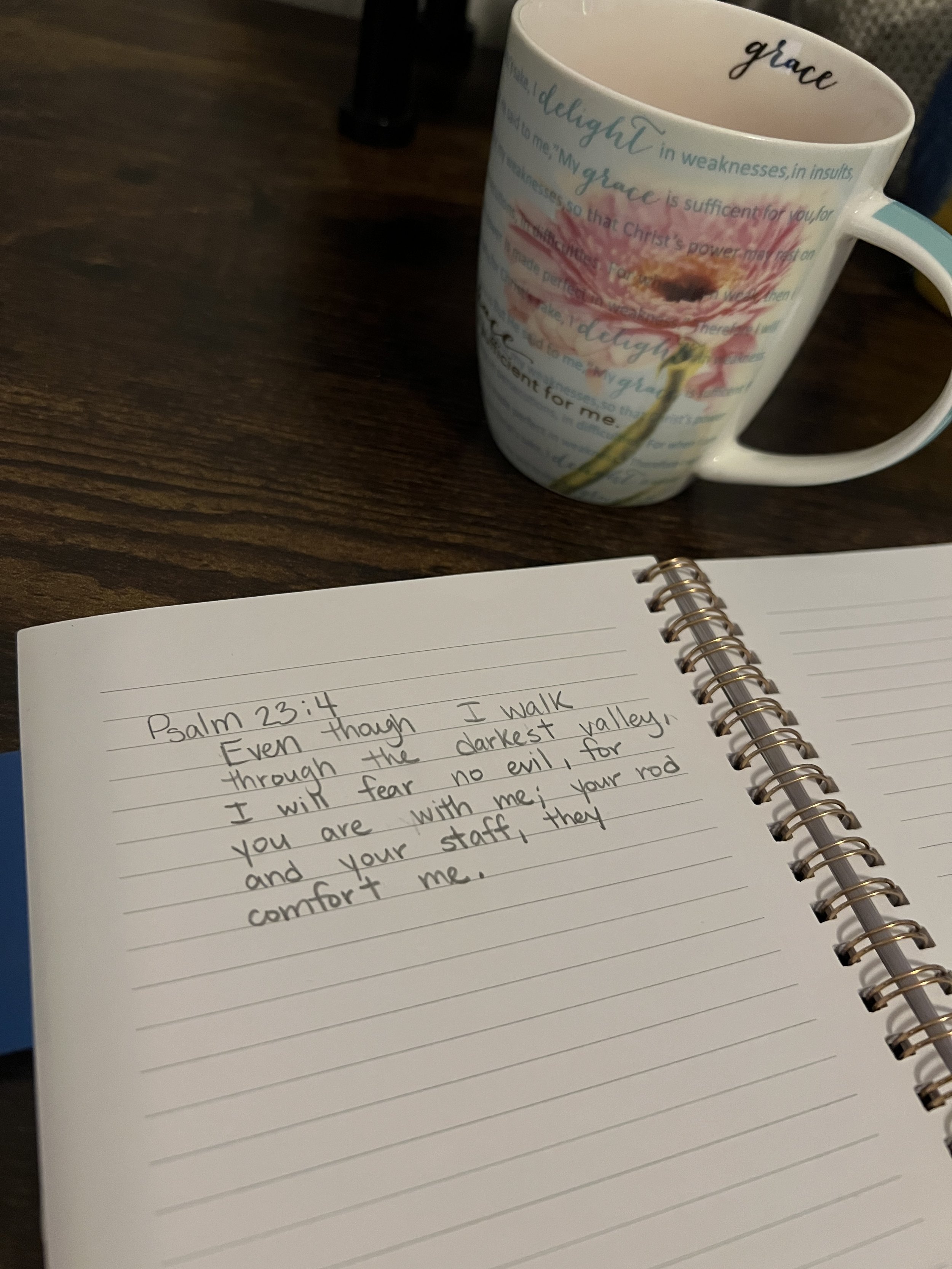 A white ceramic mug with colorful floral design and Bible scripture text, placed on a wooden surface. Next to it, an open spiral notebook with handwritten notes about Psalm 23:4.
