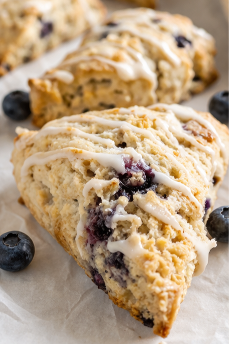 A close-up of a blueberry scone topped with white icing, with more scones and blueberries in the background.