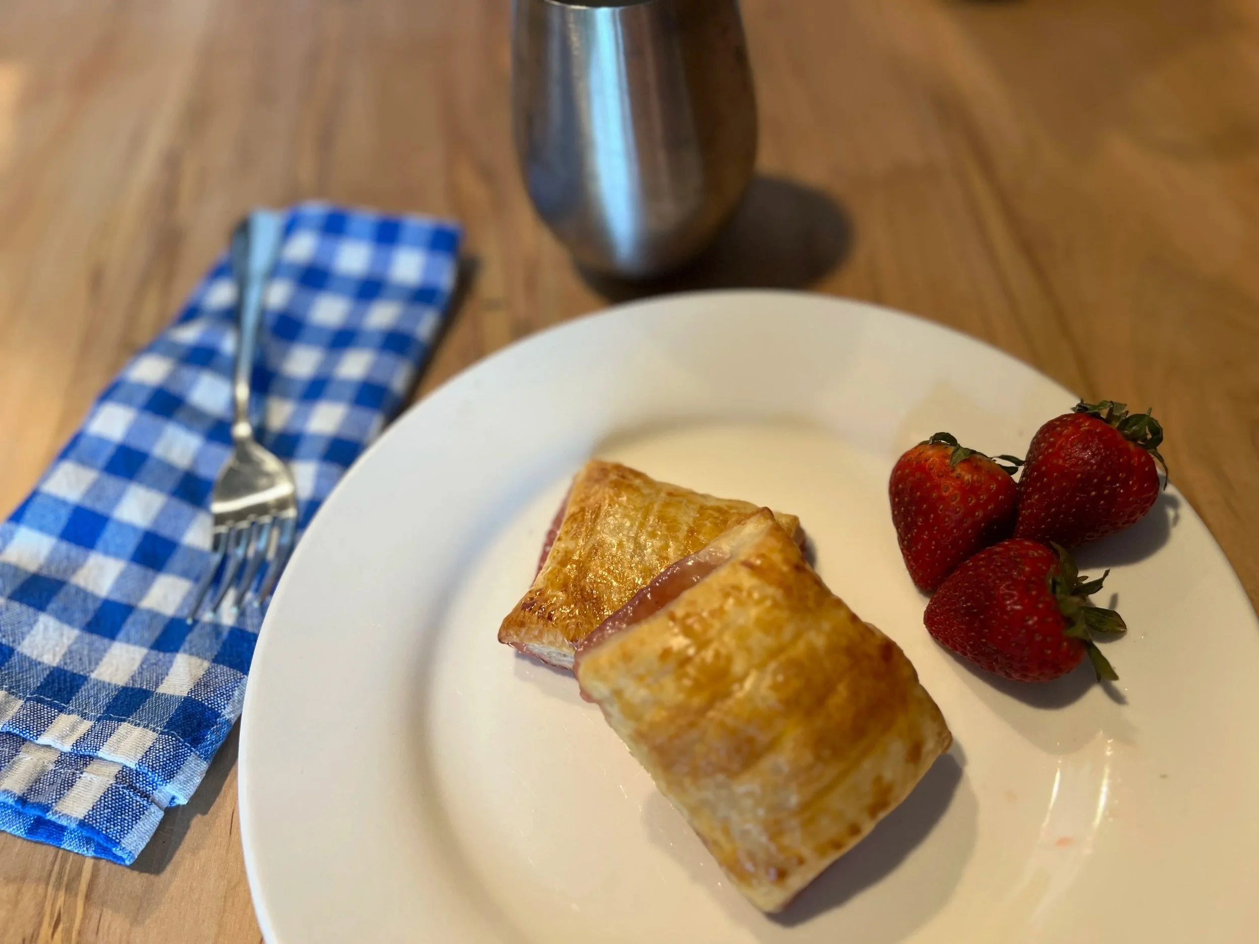 A plate with a folded pastry and fresh strawberries, on a wooden table next to a blue checkered napkin and a fork, with a metallic pitcher in the background.
