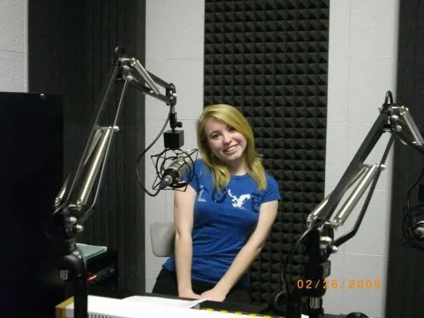A woman with blonde hair smiling and sitting in a radio or podcast recording studio, surrounded by two large microphone arms.